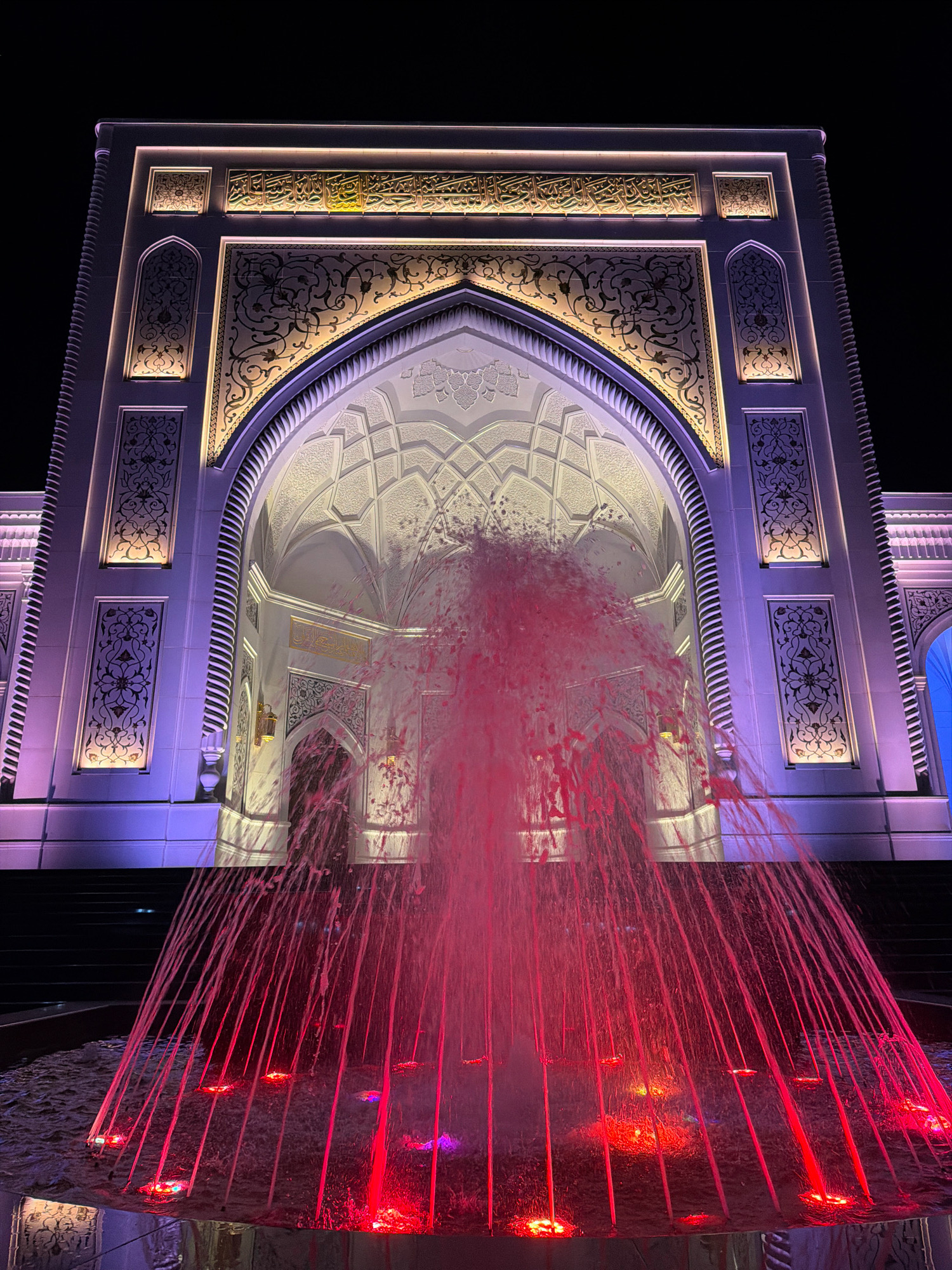 The image shows the exterior of a building, likely a mosque, at night. The building is illuminated with purple and yellow lights. The facade features intricate architectural details, including arches, decorative carvings, and calligraphy. In the foreground, a fountain sprays water illuminated by red lights. The scene appears to be in a public space, possibly a city square in Shali, Russia, given the architectural style. There are no people visible in the image.