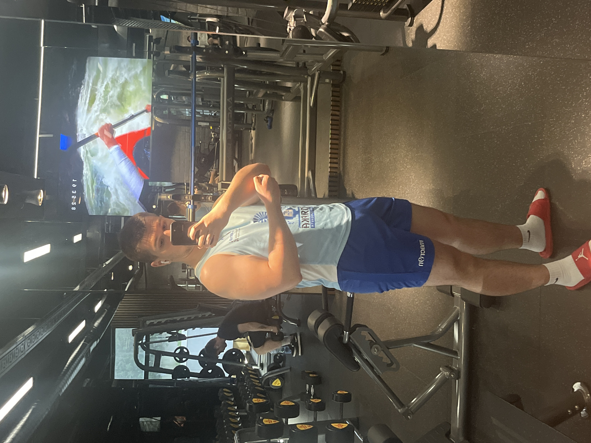 The image captures a mirror selfie taken by a young man inside a gym. The photograph is oriented 90 degrees counter-clockwise from its natural upright position.
The main subject is a young man, appearing to be in his late teens or early twenties, who is standing in front of a large mirror, holding a dark-colored smartphone in his left hand to take the photo. He is flexing his right bicep, looking towards his phone. He has short, dark hair and is wearing a light blue sleeveless tank top, blue athletic shorts, white socks with a black Nike swoosh logo, and red slide sandals featuring a white Jordan Jumpman logo.
The setting is a well-lit gym, with various pieces of exercise equipment visible in the reflection. These include racks of dumbbells, weight machines, and benches. The floor is covered with speckled, dark gym matting. In the upper left corner of the reflection, a large screen displays a video of a person kayaking in whitewater rapids, with a digital time stamp "10:32:58" visible, indicating the time of day is late morning.
Notable text details include "АКРОН FITNESS SOFIA" printed on the man's light blue tank top, which confirms the gym's location in Sofia, Bulgaria. His blue shorts also feature inverted text, possibly a brand name, but it is not clearly decipherable. The man appears focused on his pose, seemingly documenting his fitness progress or a moment during his workout.