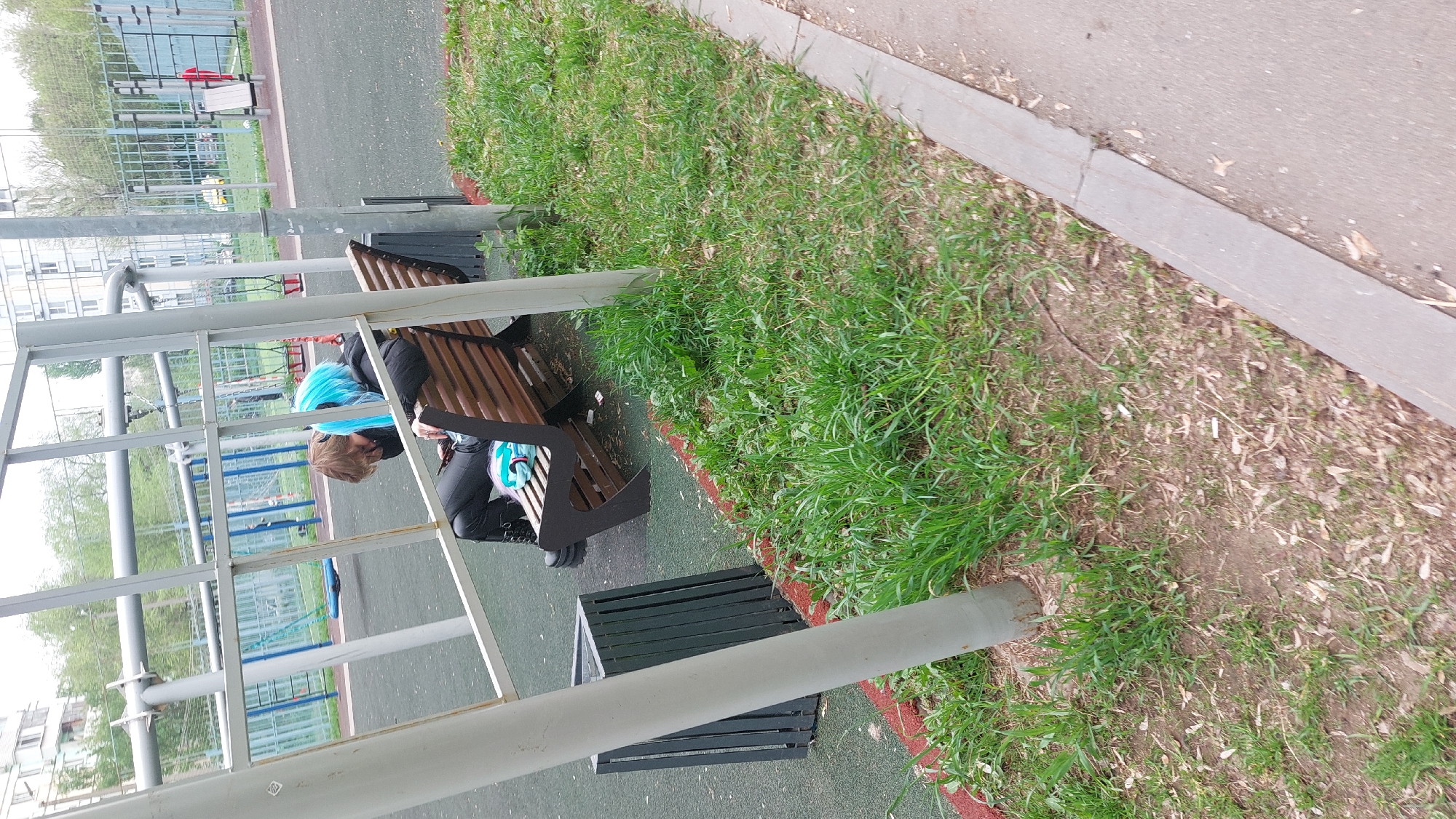 A medium-angle shot depicts an individual with blue hair resting on a wooden bench in what appears to be a park or recreational area. The bench is positioned near a grassy patch with a concrete walkway bordering it. A metal structure, possibly part of playground equipment, is visible in the foreground and adjacent to the bench. The scene is located in Moscow, Russia.