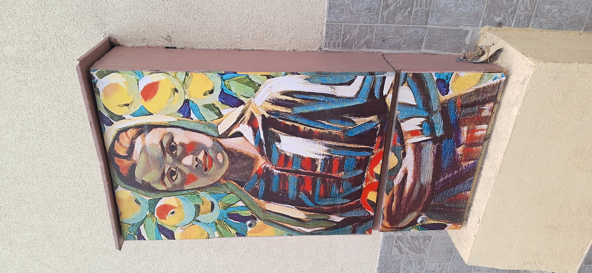 The image shows a colorful mural depicting a person lying down, surrounded by fruits, attached to the corner of a building. The mural is divided into two panels, mounted on a brown-painted structure projecting from a wall of a beige textured surface. The mural appears to be an artistic representation, with visible brushstrokes and vibrant colors. Adjacent to the mural are sections of the building's facade, including a light-colored wall and a tiled area of gray and white. Based on the prompt, this mural is located in Blagoevgrad, Bulgaria.