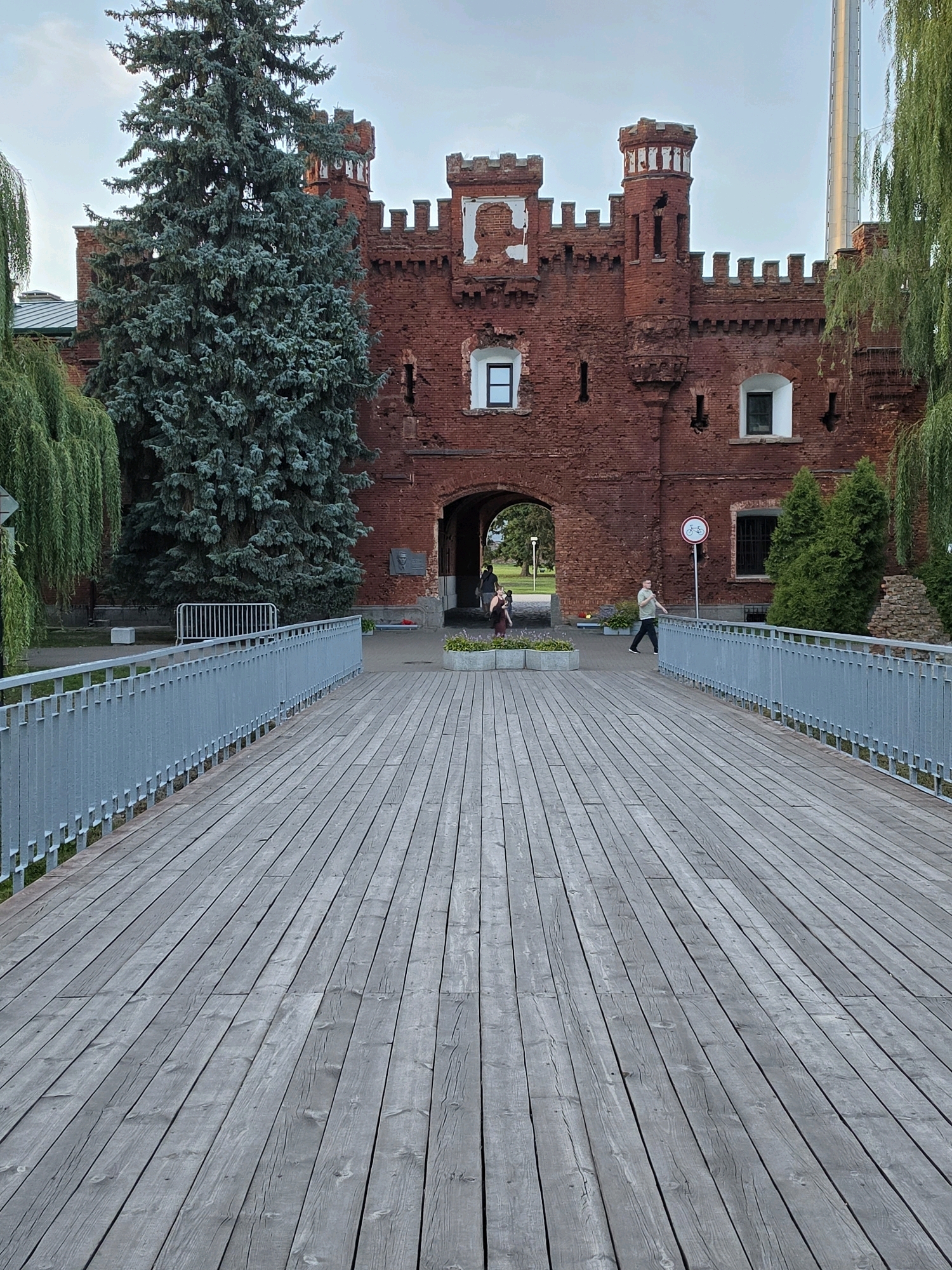 The image shows the entrance to the Brest Fortress in Brest, Belarus. The fortress is made of red brick and has a crenelated design with turrets. In the foreground, there is a wooden walkway with a metal railing leading to the fortress entrance. Several people are visible in the image. Two individuals are walking through the archway of the fortress, and one person is walking on the walkway towards the fortress. A large evergreen tree stands to the left of the entrance, and weeping willow trees are visible on either side of the walkway. A bicycle-restricted zone sign is posted on the right side of the walkway. The sky is overcast.