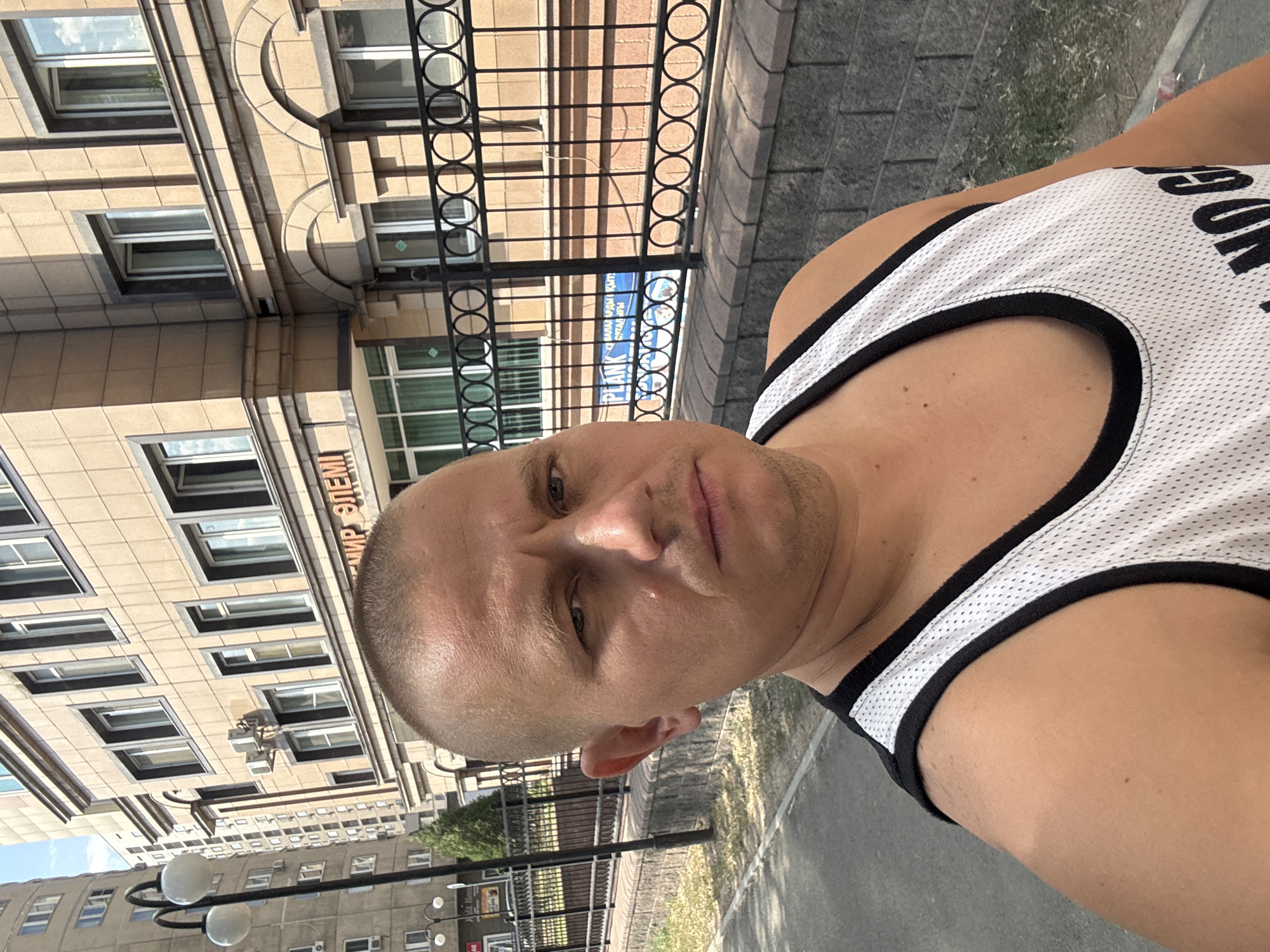 A man with a shaved head and a serious expression takes a selfie outdoors, with a building and street in the background. He is wearing a white tank top with black trim. The building behind him is a multi-story structure with beige siding and numerous windows, some of which are open. A black metal fence runs along the front of the building. A street light with two globe-shaped lamps is visible to the left of the man. A sidewalk and grassy area are also present in the foreground. The scene appears to be in an urban environment. Based on the text visible on the building which reads "МИР ЭЛЕМ" (MIR ELEM), this location is likely in Almaty, Kazakhstan, as this is a common business name there.