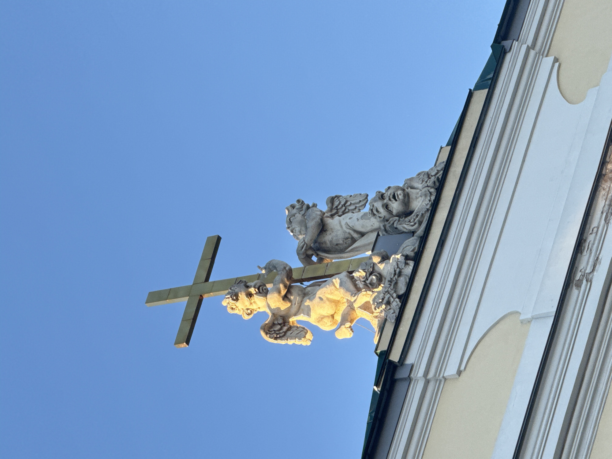The image shows a low-angle view of a building's exterior. A golden cross is held by stone angel statues. The building has architectural details with white trim and beige walls. The background is a clear blue sky. The building is located in Saint Petersburg, Russia.