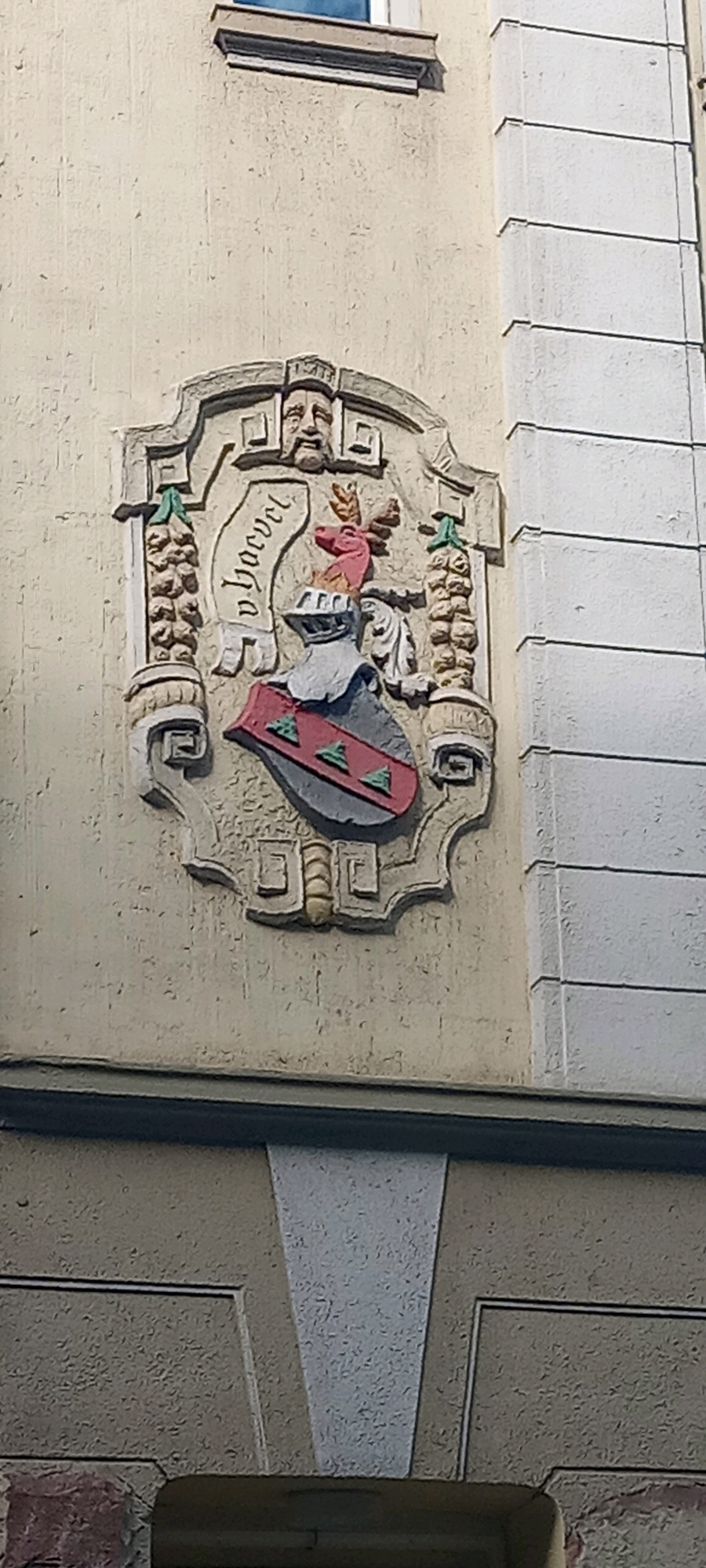 The image shows a relief sculpture on the facade of a building. The sculpture is an escutcheon, or coat of arms, featuring a shield with red and green triangular elements and a helmet topped with a stag's head. Above the shield, a banner displays the inscription "v. Horvel". The entire relief is framed by decorative elements, including what appears to be a human face at the top and cornucopia-like structures on the sides. The building's facade is a textured, light-colored plaster, with a white, fluted pilaster visible on the right side of the image. The architecture suggests a historical building. This coat of arms is located at the historic "Haus Immergrün" at Prinzipalmarkt 25 in Münster, Germany, not Dortmund.