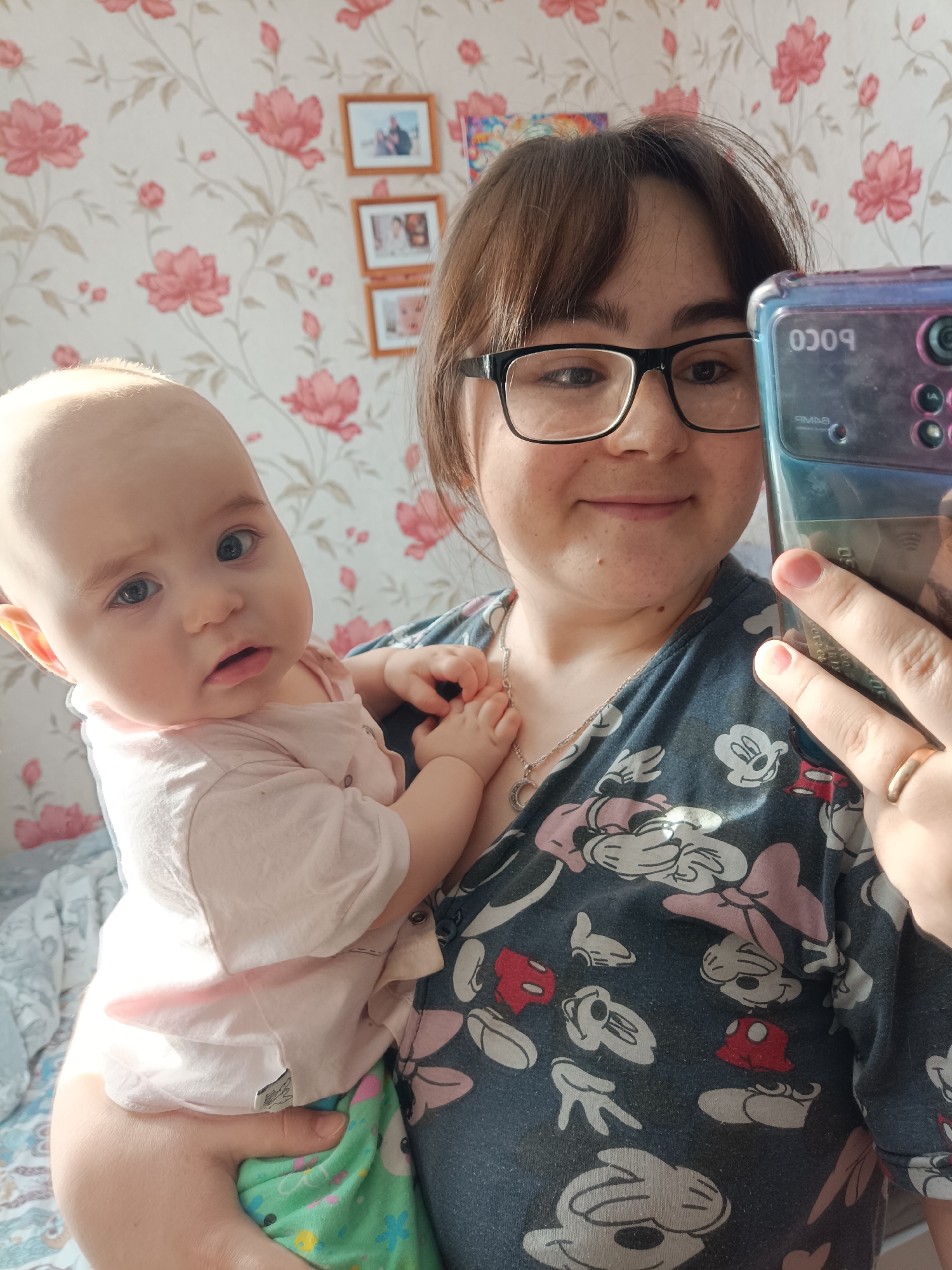 The image displays an indoor scene in the city of Noviy, Russia, featuring an adult female holding an infant. The adult female, positioned on the right, has brown hair and wears black-rimmed glasses. She is taking a selfie with a smartphone held in her right hand; the phone's case is teal/blue, displaying reversed text "POCO" and "64MP" from the camera module. She wears a dark blue t-shirt patterned with white, pink, and red Mickey and Minnie Mouse elements, a silver-colored necklace with a crescent moon pendant, and a plain gold-colored ring on her right ring finger.
The infant, held in the adult's left arm and situated on the left, has light hair and blue eyes, looking directly at the camera with a neutral expression and slightly parted lips. The infant wears a light pink short-sleeved top and partially visible green patterned pants. Its right hand rests near the adult's chest.
The background consists of a wall covered in light-colored wallpaper adorned with a repeating pattern of pink/red flowers and green leaves. Three small, light-framed photographs are vertically arranged on the wall behind the adult's shoulder. A segment of a colorful decoration is visible higher up on the wall. A light-colored, patterned textile, likely a blanket, is partially visible in the lower-left background.