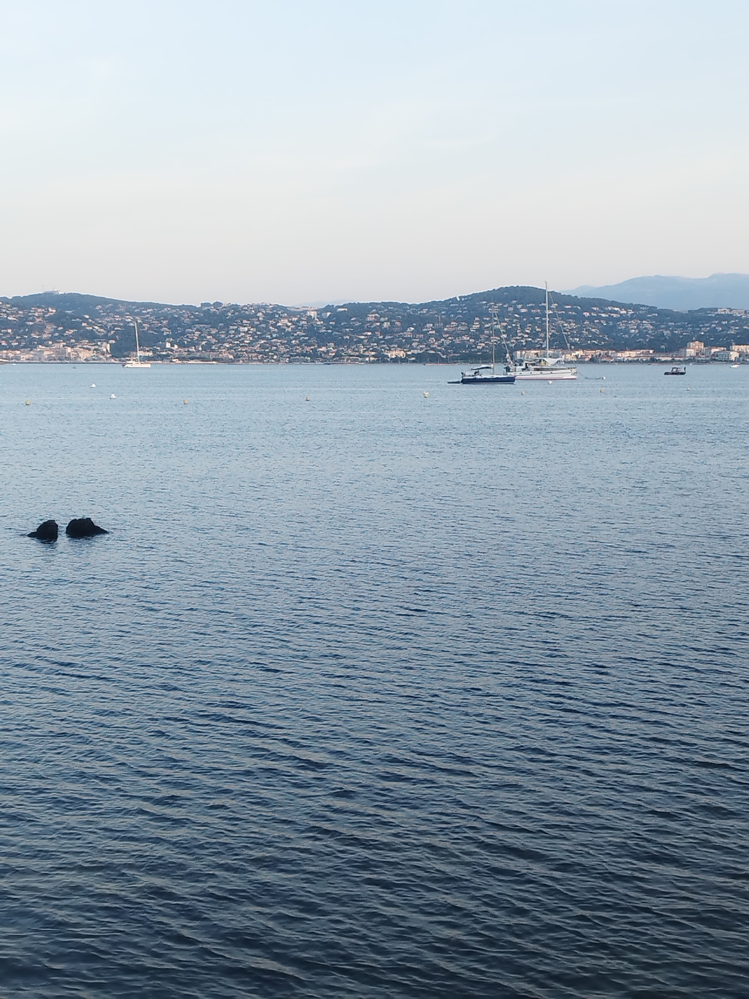 The image depicts a tranquil scene of the sea in front of Antibes, France. The water is calm with gentle ripples, reflecting the soft, clear sky. In the distance, a picturesque coastline is visible, dotted with numerous buildings that form the cityscape of Antibes. The shoreline is backed by rolling hills covered in vegetation. Several boats are present on the water, including sailboats and a smaller motorboat, adding a sense of activity to the otherwise serene environment. Closer to the foreground, two dark rocks emerge from the water. The overall impression is one of peaceful coastal living, with the natural beauty of the sea complementing the urban landscape.