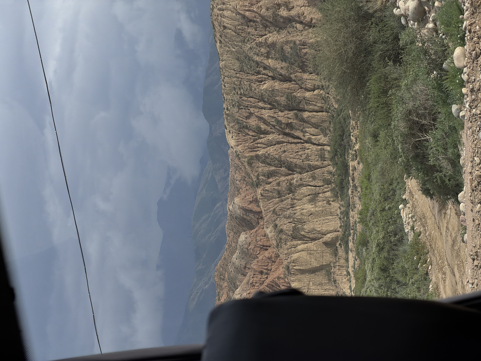 The image depicts a vast, rugged landscape under a cloudy sky, likely taken from within a vehicle, as evidenced by the dark interior element in the foreground and a visible wire across the top portion. The prominent feature is a series of eroded hills and mountains exhibiting distinct layers of earthy tones, ranging from light brown to reddish hues. These formations show deep vertical striations, indicative of significant weathering and erosion. Sparse green vegetation, including bushes and grasses, dots the slopes and the valley floor. A dirt road with tire tracks winds through the lower part of the scene, suggesting human presence or activity in the area. In the background, more distant mountains fade into a hazy, overcast sky. The overall impression is one of a remote, natural environment with dramatic geological features. The context provided by the city of Tong, Kyrgyzstan, points to this being a scenic area within that region.