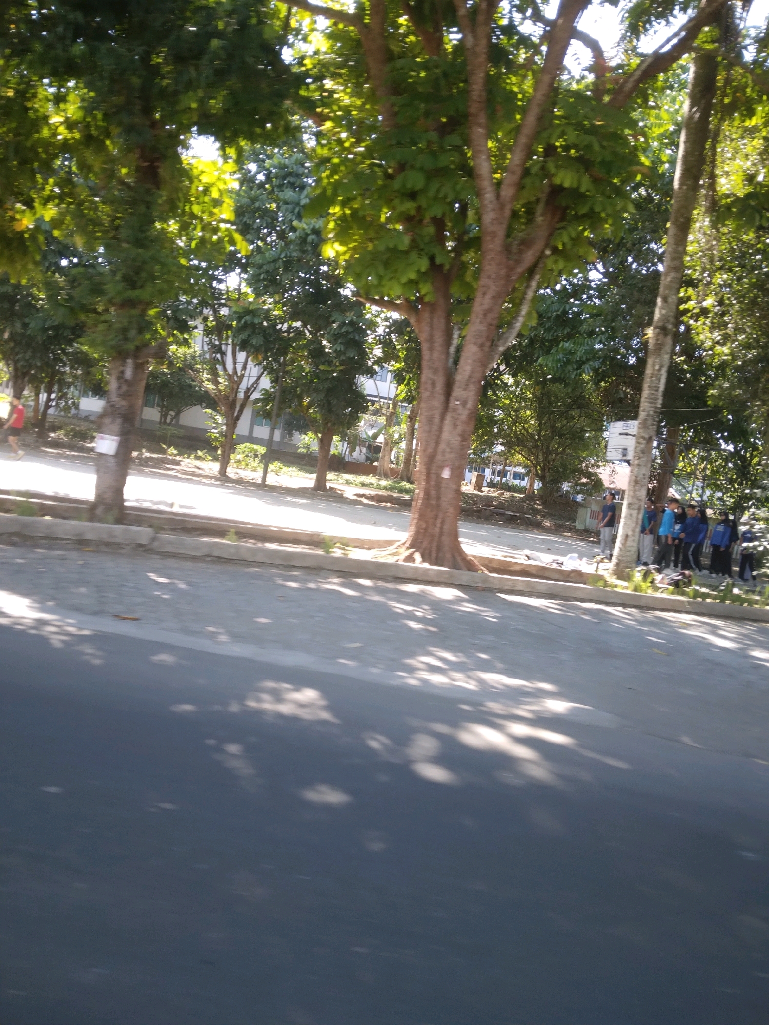 The image depicts a street scene in Grendeng, Indonesia, on a sunny day. A line of people, appearing to be students, stands on the side of the road, some in uniform. Trees with green foliage line the street, casting shadows on the asphalt. A white building is visible in the background. The scene suggests a typical day in an urban or semi-urban environment in Indonesia.