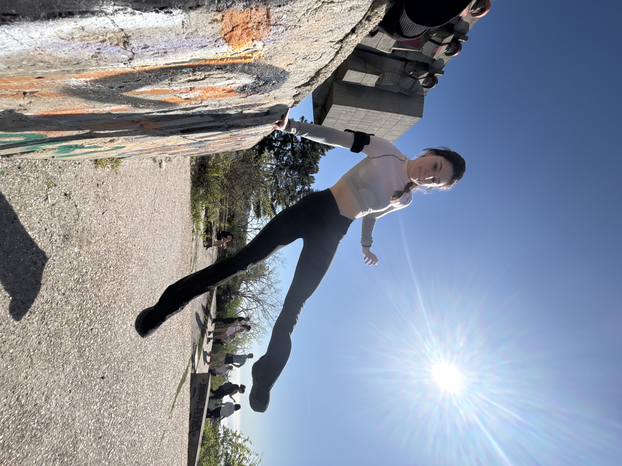 A girl hangs from a concrete structure. She is wearing a long sleeve white shirt and black leggings. Her hair is in a braid. The concrete structure has graffiti. A crowd of people stands at the bottom. The sky is blue. The sun is shining brightly.