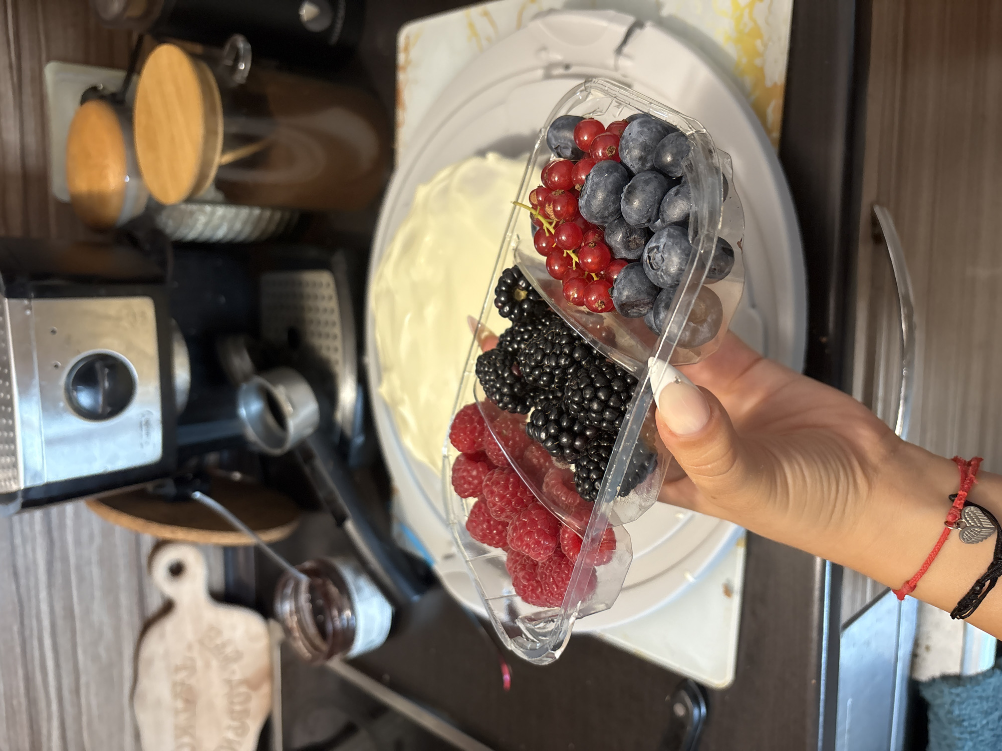 An overhead medium shot captures a hand holding a clear plastic container of fresh berries, including raspberries, blackberries, blueberries, and red currants. Behind the berries, a white frosted cake is visible on a white cake stand. The background features kitchen appliances, including a silver espresso machine, and two glass jars with wooden lids containing dark contents. A small jar of jam and a wooden cutting board are also present. The scene appears to be set in a domestic kitchen environment.