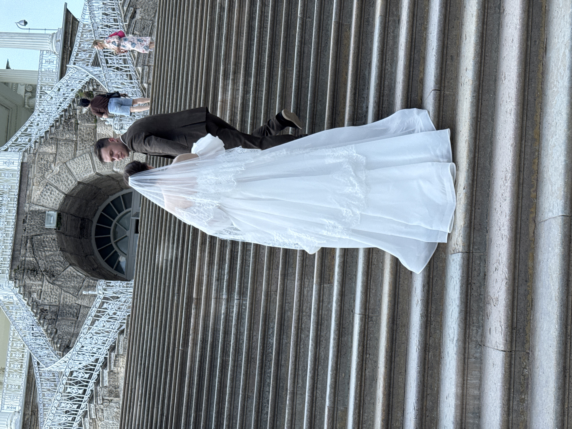 A wedding couple is ascending a wide, stone staircase. The bride, seen from behind, wears a traditional white wedding gown with a veil and is positioned slightly ahead of the groom, who is in a dark suit. Two other individuals, one in a brown shirt and denim shorts, and another in a floral dress, are also on the stairs further up. The staircase leads to a building with a large arched window and ornate white railings on either side. The scene is set in front of the Chinese Grand Palace in the city of Pushkin, Russia, part of the Tsarskoye Selo complex.