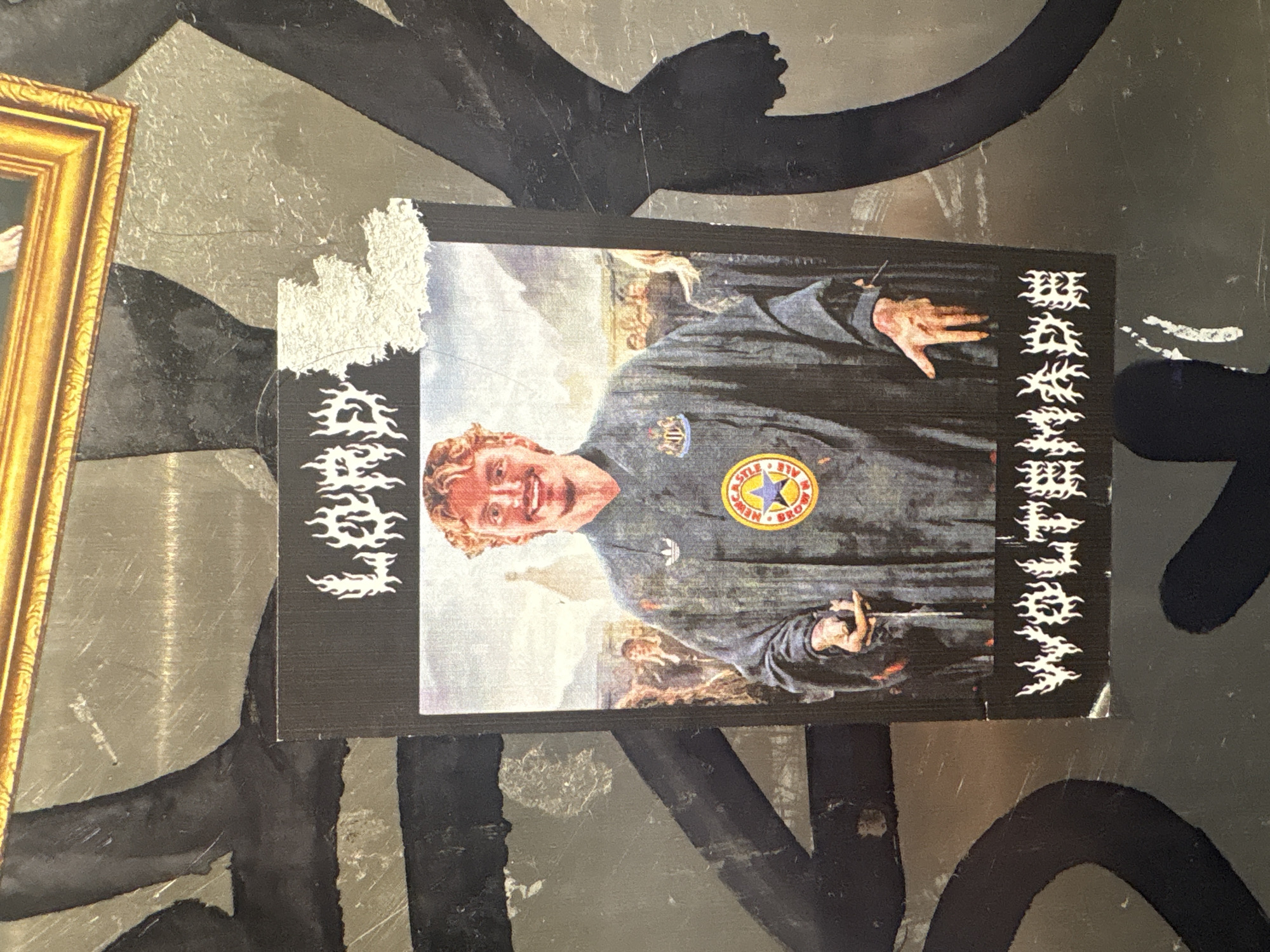 A sticker depicting an image of a man in a blue garment is partially adhered to a textured surface. The man is wearing a garment with a Newcastle Brown Ale logo. The background behind the figure is a classical artwork depicting a scene of multiple figures and light sky. The sticker has decorative text running along the sides of the sticker. It is located in London, United Kingdom, specifically the surface it is adhered to is adjacent to a gold picture frame.