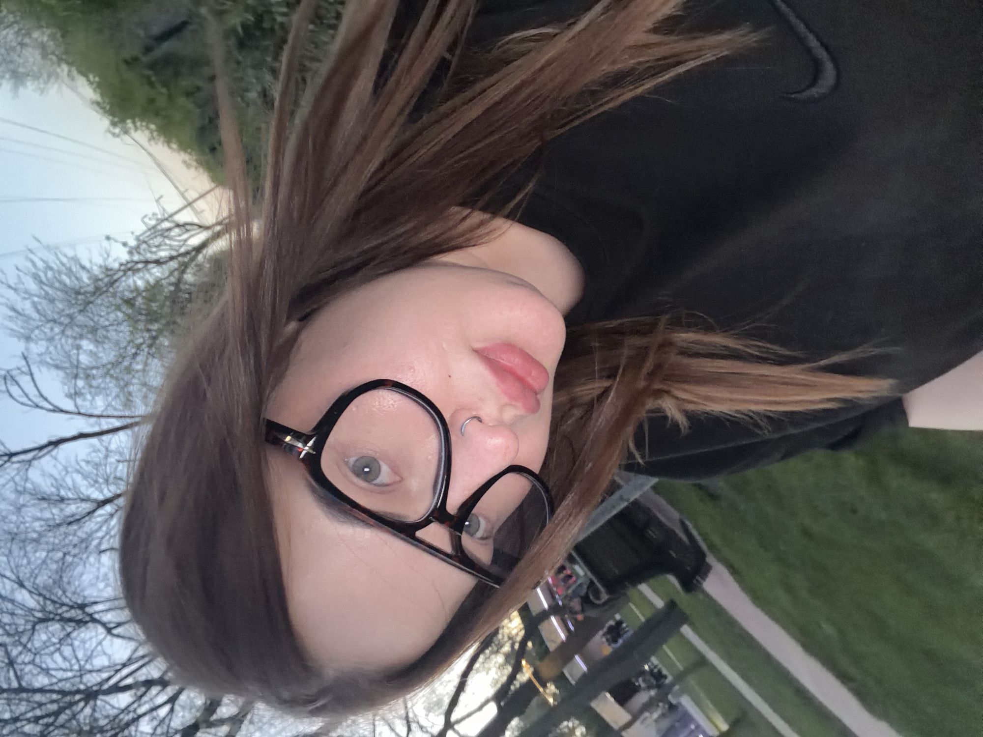 A young woman with brown hair and glasses wearing a black sweatshirt is photographed outdoors. She has a nose ring in her left nostril. The background includes trees without leaves, suggesting late fall or early spring, and a green grassy area with what appears to be a sidewalk and possibly some structures or vehicles in the distance. The location cannot be determined to be in Krasnodar, Russia based on this image alone.