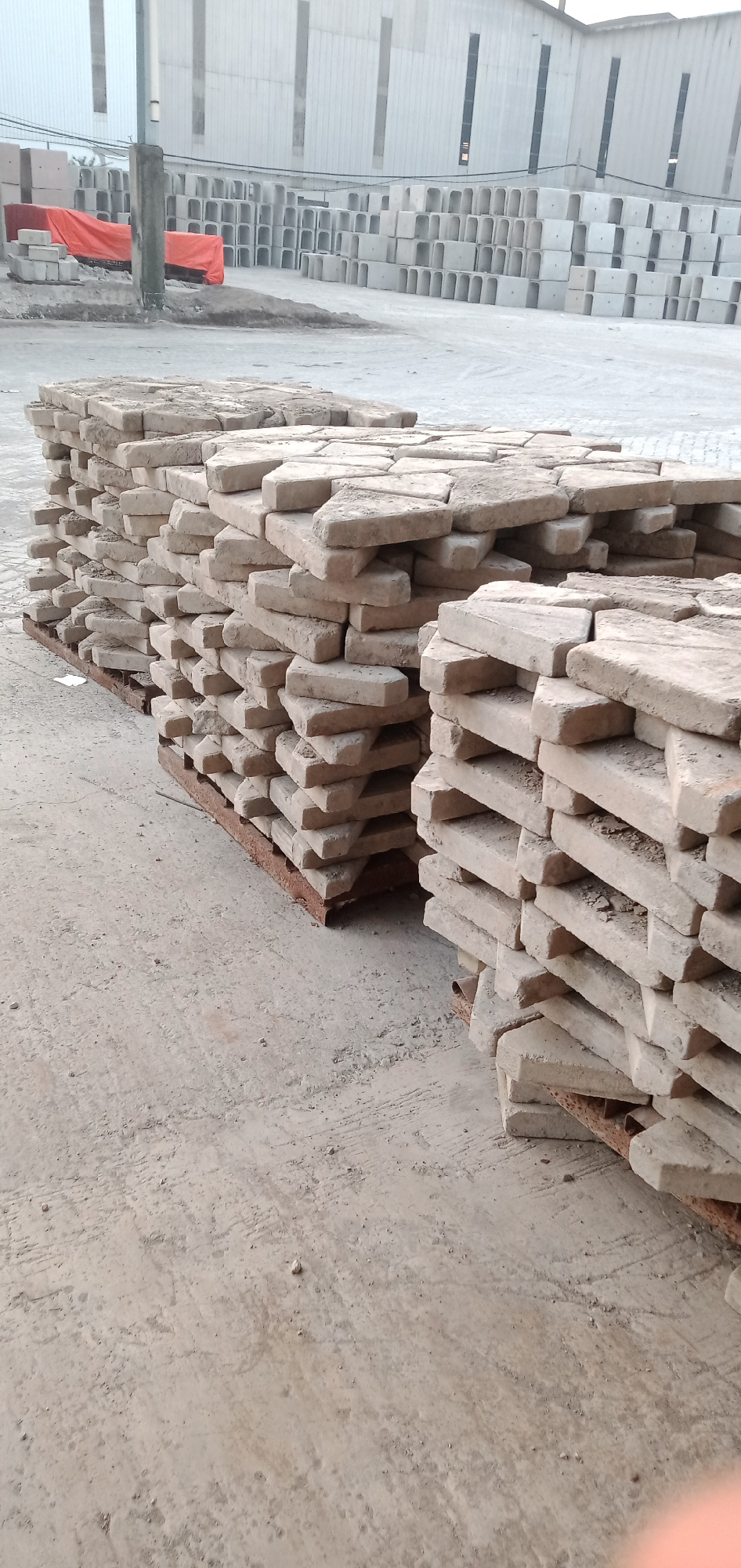 The image shows stacks of rectangular paving stones on wooden pallets. The stones are light beige and appear dusty or weathered. In the background, there is a large building with tall, narrow windows and stacks of U-shaped concrete structures. An orange tarp partially covers some of the structures in the background. The location is Sumput, Indonesia, likely an industrial or storage area. There are no people visible. The focus is on the paving stones and the materials yard environment.