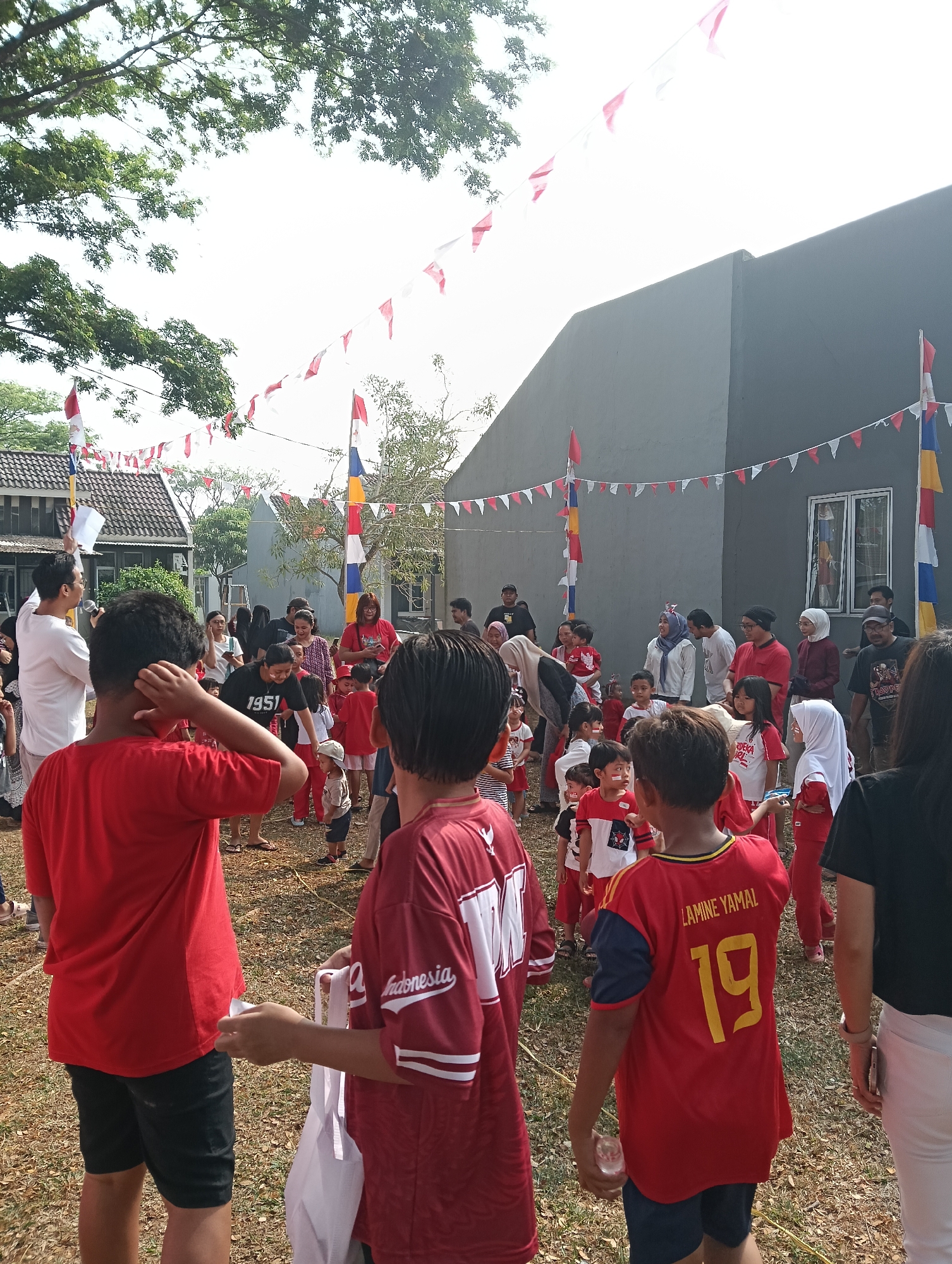 In Parung Panjang, Indonesia, a group of people, primarily children and adults, are gathered outdoors in a grassy area. Many are wearing red and white clothing, and there are red and white festive banners strung across the scene, suggesting a celebration or event. Some children have face paint on. In the foreground, a young boy in a red shirt with the name "LAMINE YAMAL" and the number "19" on his back is visible. To his left, another boy in a red shirt is holding a white plastic bag. Adults are observing the children and participating in activities. A grey building with a window is in the background, and trees are visible overhead. The overall atmosphere appears to be lively and festive.