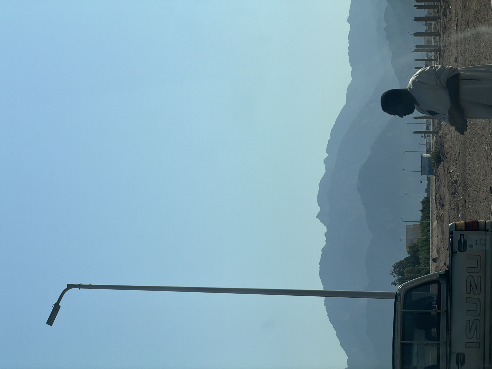 In Dahab, Egypt, a man stands in profile, facing left, his hands clasped in front of him. He wears a light-colored, long-sleeved garment. The background features a vast, clear blue sky, and a silhouetted mountain range in the distance. In the foreground, a silver pickup truck with "ISUZU" visible on its tailgate is parked. A tall streetlamp stands to the left of the frame. The environment appears to be arid, with sandy terrain and scattered construction elements, including wooden poles.