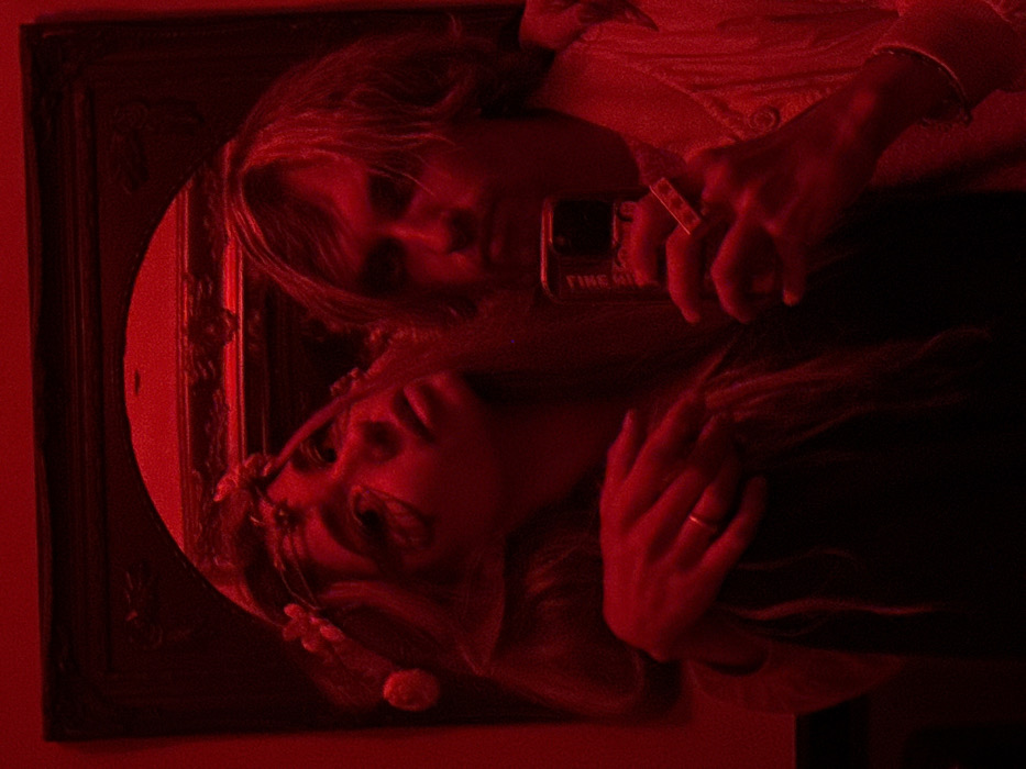 This image, taken in the city of Tel-Aviv, Israel, displays a mirror selfie of two individuals bathed in a pervasive red light. A female subject is positioned on the left side of the frame, with long, light-colored hair, a floral headband, and prominent eye makeup. Her right arm is draped over the shoulder of the male subject to her right, and a gold-colored ring is visible on her left ring finger. The male subject, with medium-length light-colored hair, holds a dark-colored smartphone in his left hand, oriented to capture the reflection. He holds a lit cigarette between the index and middle fingers of his right hand, which also features a silver-colored bracelet on his wrist. He wears a light-colored collared shirt. Both individuals appear to be looking towards the camera/mirror. The subjects are reflected in an ornate, dark-framed mirror, suggesting an indoor setting.