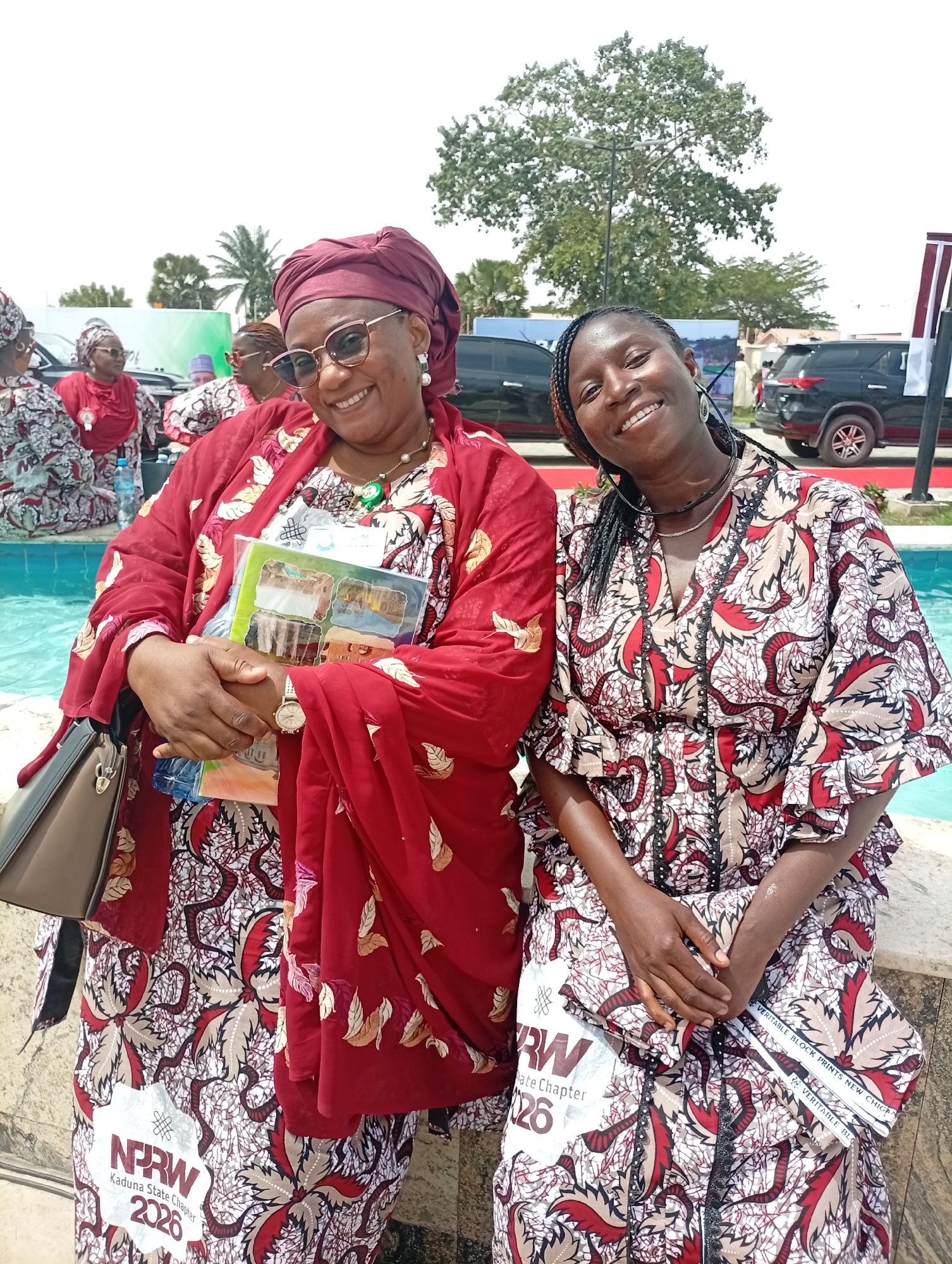 Two women are prominently featured in a bright outdoor photograph, likely taken at an event in Unguwan Sanusi, Nigeria. The woman on the left is smiling broadly, wearing sunglasses, a red headwrap, and a flowing red kaftan adorned with gold leaf-like patterns. She holds a stack of books or documents with colorful covers and wears a patterned dress underneath her kaftan. Her accessories include a necklace, earrings, and a wristwatch, with a dark handbag visible on her left.
Beside her, the woman on the right is also smiling, dressed in a garment with a matching red, beige, and black patterned fabric, which appears to be the same textile as the inner dress of the woman on the left. She has braided hair and wears necklaces and large hoop earrings. Both women are seated next to a blue-tiled water feature, possibly a fountain or pool.
In the background, other women in similar attire can be seen, along with dark SUVs, lush green trees, and distant structures, indicating an outdoor gathering or event. The overall atmosphere appears joyful and social, conveyed by the subjects' expressions.
Visible text on the patterned fabric includes "NPRW Kaduna State Chapter 2026" and a textile brand label that reads "VERITABLE BLOCK PRINTS NEW CHIGAN". These details suggest the event is associated with the "NPRW Kaduna State Chapter" and that the traditional attire is made from a specific type of printed fabric.