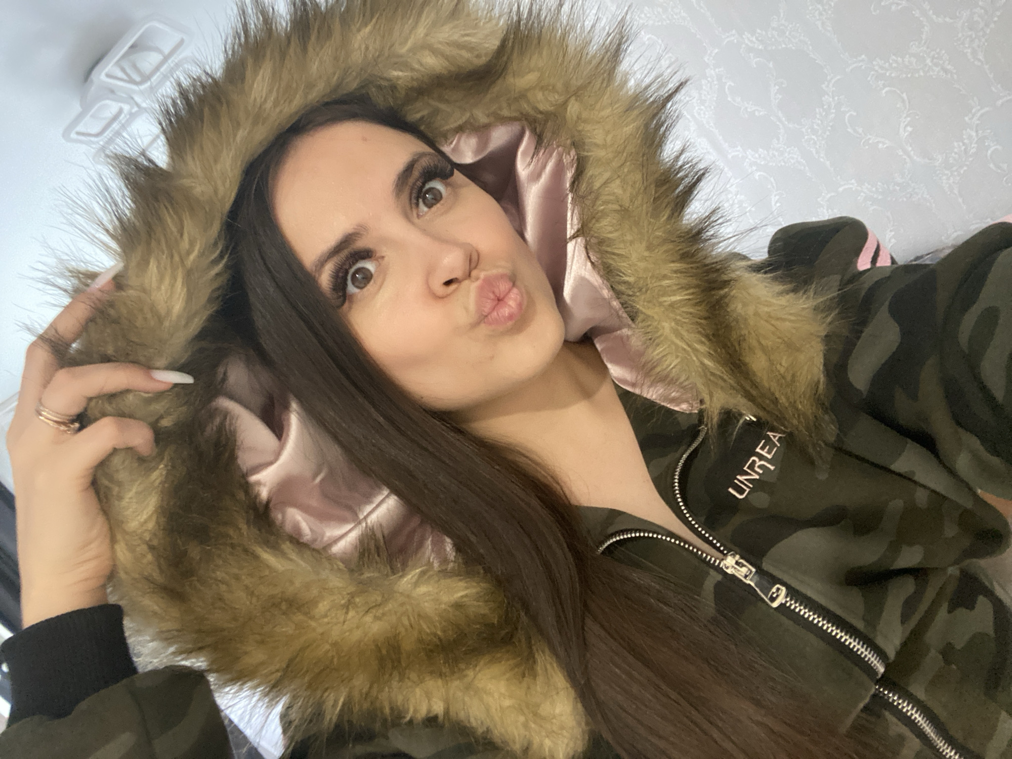 A woman is posing for a selfie, wearing a camouflage jacket with a fur-lined hood. The jacket has a pink lining, and the word "UNREA" is visible on the upper left. Her hair is long, brown, and straight. She is puckering her lips in a kiss face. Her eyes are wide and looking upwards. She is holding the fur trim of the hood with her left hand, showing long nails. There is a ring on her left ring finger. The background is a white wall with a subtle patterned wallpaper. The location in the city of Chaikovsky, Russia cannot be determined from the image.