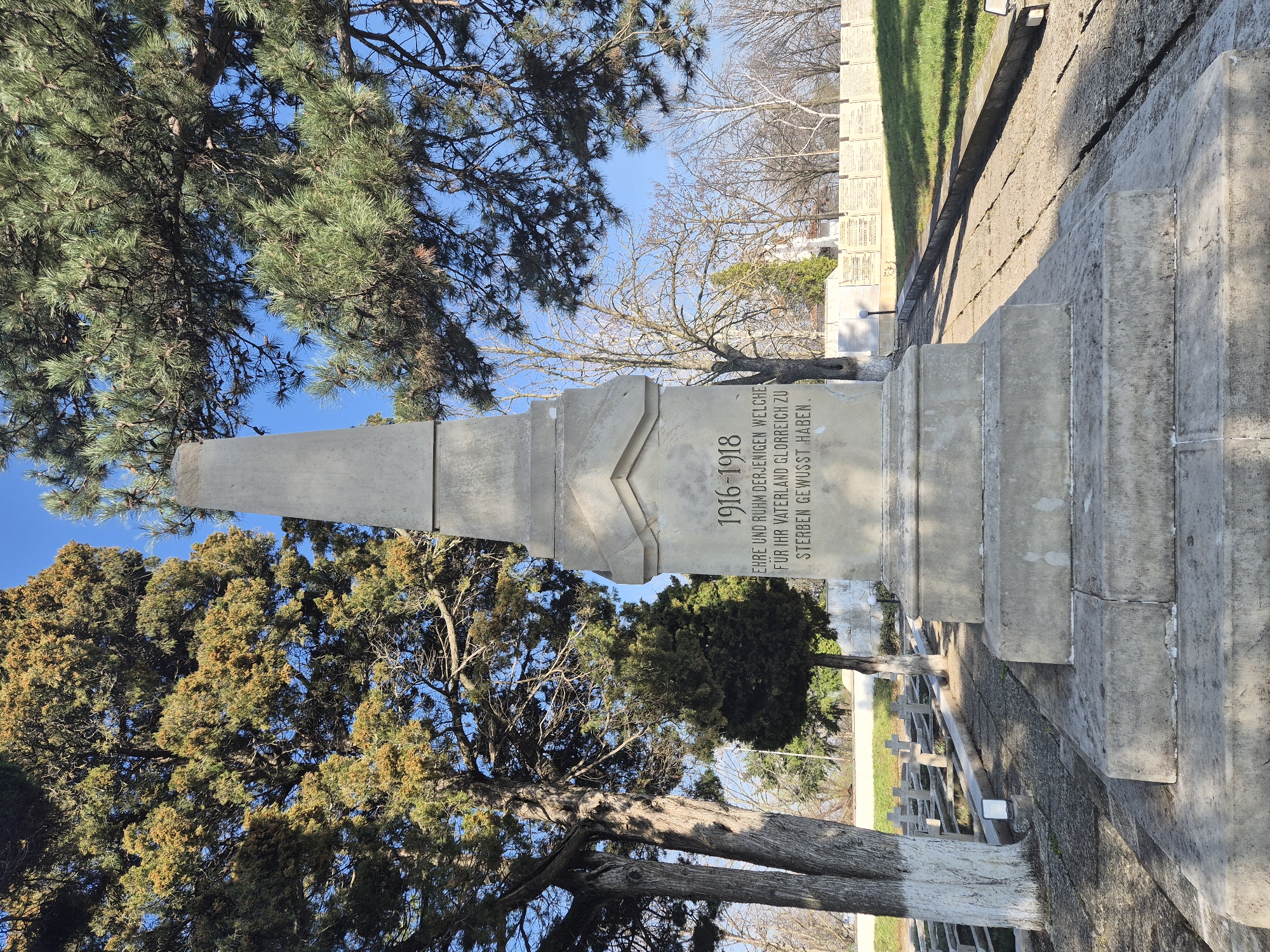 The image depicts a World War I memorial in a cemetery setting. The inscription reads "1916-1918, EHRE UND RUHM DERJENIGEN WELCHE FUR IHR VATERLAND GLORREICH ZU STERBEN GEWUSST HABEN." which translates to "1916-1918, HONOR AND GLORY TO THOSE WHO KNEW HOW TO DIE GLORIOUSLY FOR THEIR FATHERLAND." The memorial is made of stone and stands amidst trees and other grave markers. It is located in Shumen, Bulgaria.