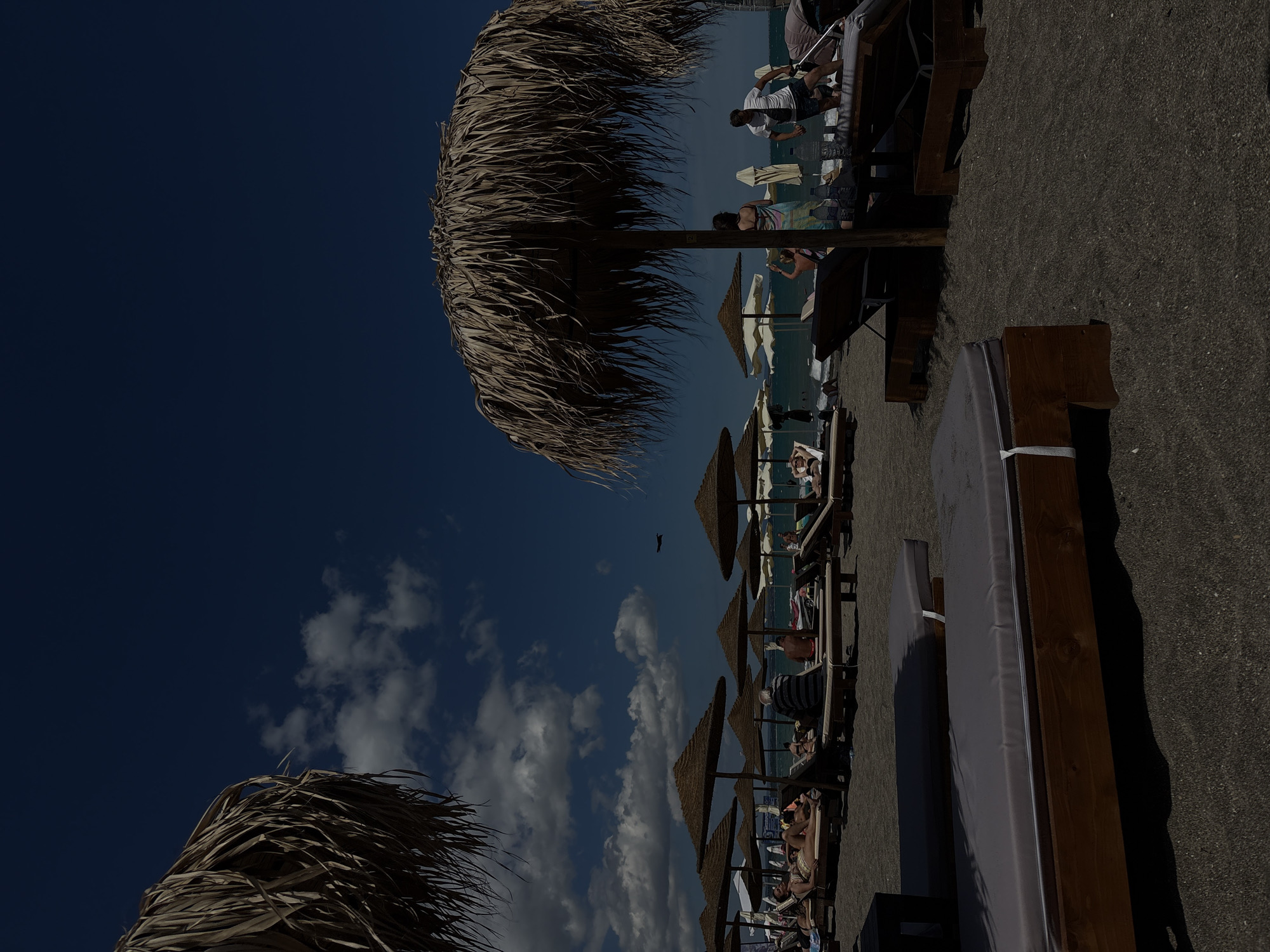 The image depicts a beach scene in Burgas, Bulgaria, under a clear blue sky with scattered white clouds. Several beach umbrellas made of dried palm leaves provide shade for rows of lounge chairs. People are seen relaxing on the lounge chairs, some sunbathing, while others appear to be walking or interacting. The foreground features two empty wooden lounge chairs with gray cushions, positioned on dark sand. In the mid-ground, more lounge chairs and umbrellas are visible, occupied by beachgoers. The sea is visible in the background, with a gentle wave pattern. A small airplane is seen in the sky. The overall atmosphere is one of a typical summer day at the beach.