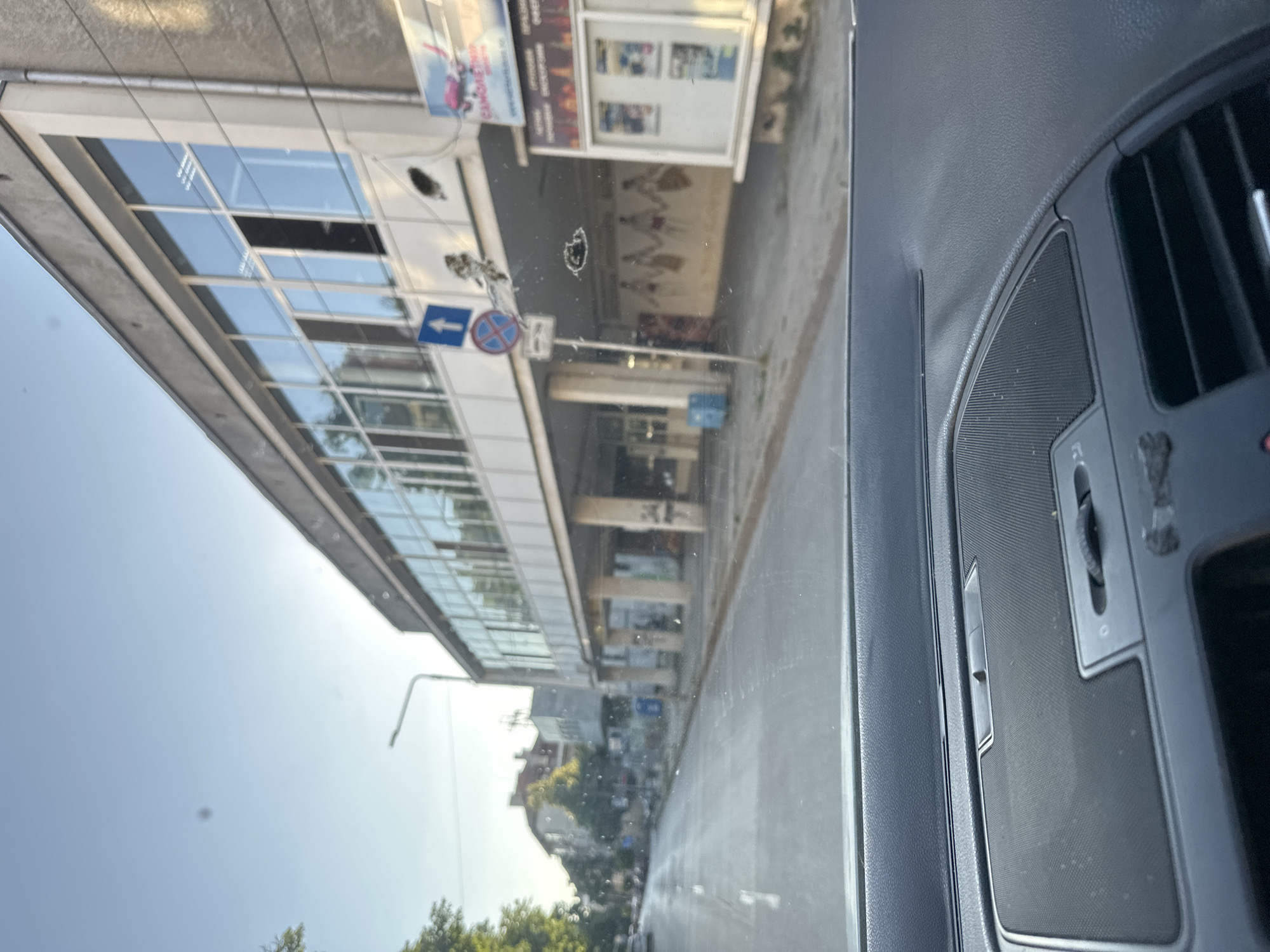 The image, taken from inside a vehicle, depicts a street scene in Ruse, Bulgaria. The view is directed forward, capturing a road with faint lane markings and parked cars visible in the distance on the left. Dominating the right side of the frame is a multi-story building with a modern facade featuring large, reflective windows. Below the windows, the building has an open ground floor supported by columns. On the exterior of this building, there are several signs and a mural of figures holding hands. A street light and traffic signs are also visible. The sky above is clear and blue, suggesting daylight. The interior of the vehicle is partially visible in the foreground, showing a dashboard with a speaker grille and air vents.