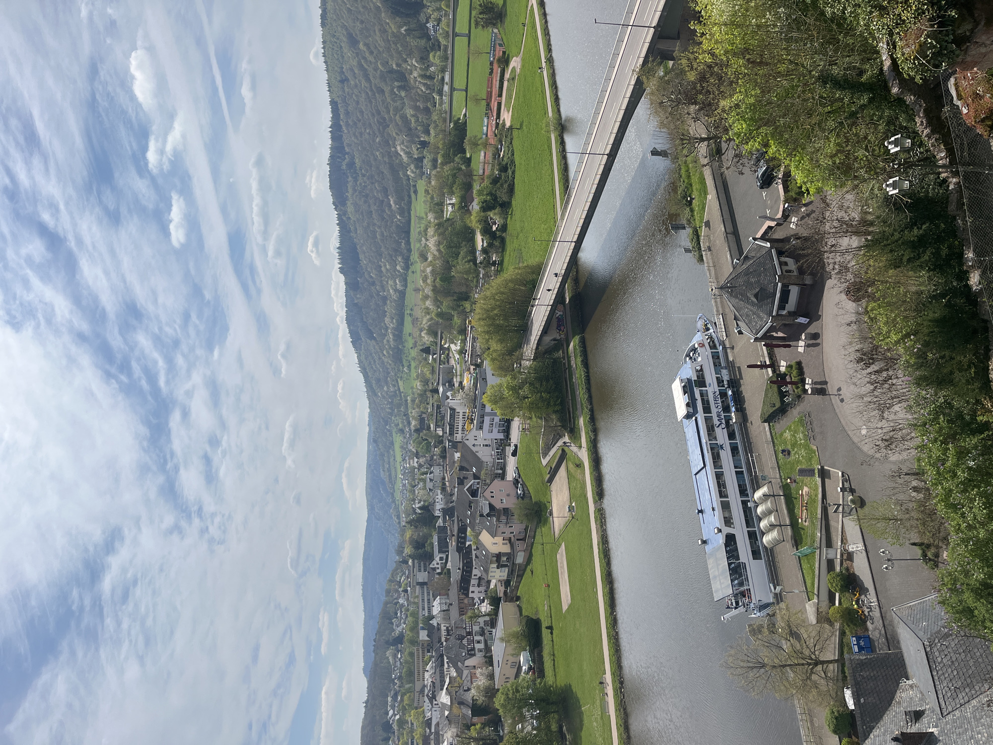 The image is a high-angle, slightly tilted view of a river and a town, likely Saarburg, Germany, based on the identifiable landmarks. The river runs vertically through the center, with a long, modern passenger ship docked along the right bank. A bridge spans the river in the upper portion of the image. The right side of the image includes a paved walkway along the river, with streetlights and trees lining the path. A building with a dark roof is also visible. The left side of the river displays the town, comprised of densely packed buildings with varying roof colors, predominantly red and gray. Beyond the town, a hillside rises, covered in green vegetation and trees. The sky above is partially cloudy with wispy cirrus formations.