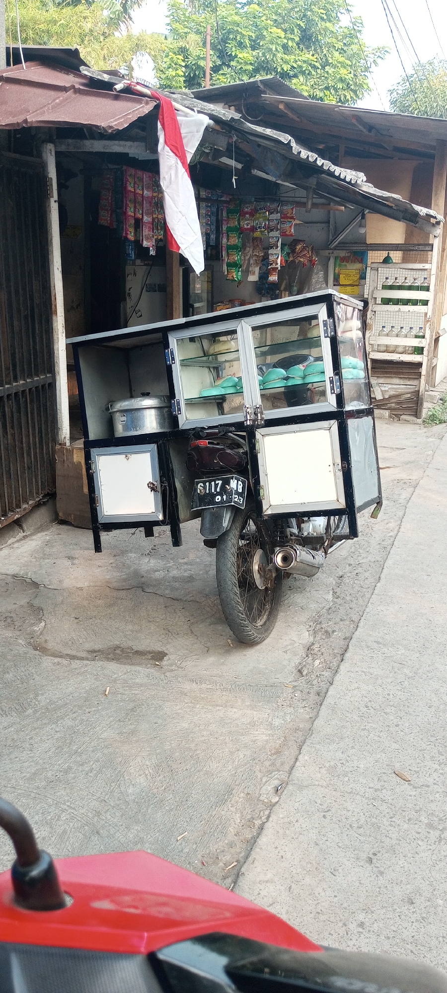 A red and white Indonesian flag is displayed outside a small, open-air store selling snacks. The store is constructed from corrugated metal and wood, with a variety of packaged goods arranged on shelves. In front of the store, a motorcycle is parked, modified with a display case attached to its rear. The case is made of glass and metal and holds several teal-colored food items. The motorcycle has a license plate that reads "B 6117 MB 01-23". The ground is paved with concrete, showing some cracks and debris. The scene is located on a street in Setu, Indonesia, evidenced by the license plate format.