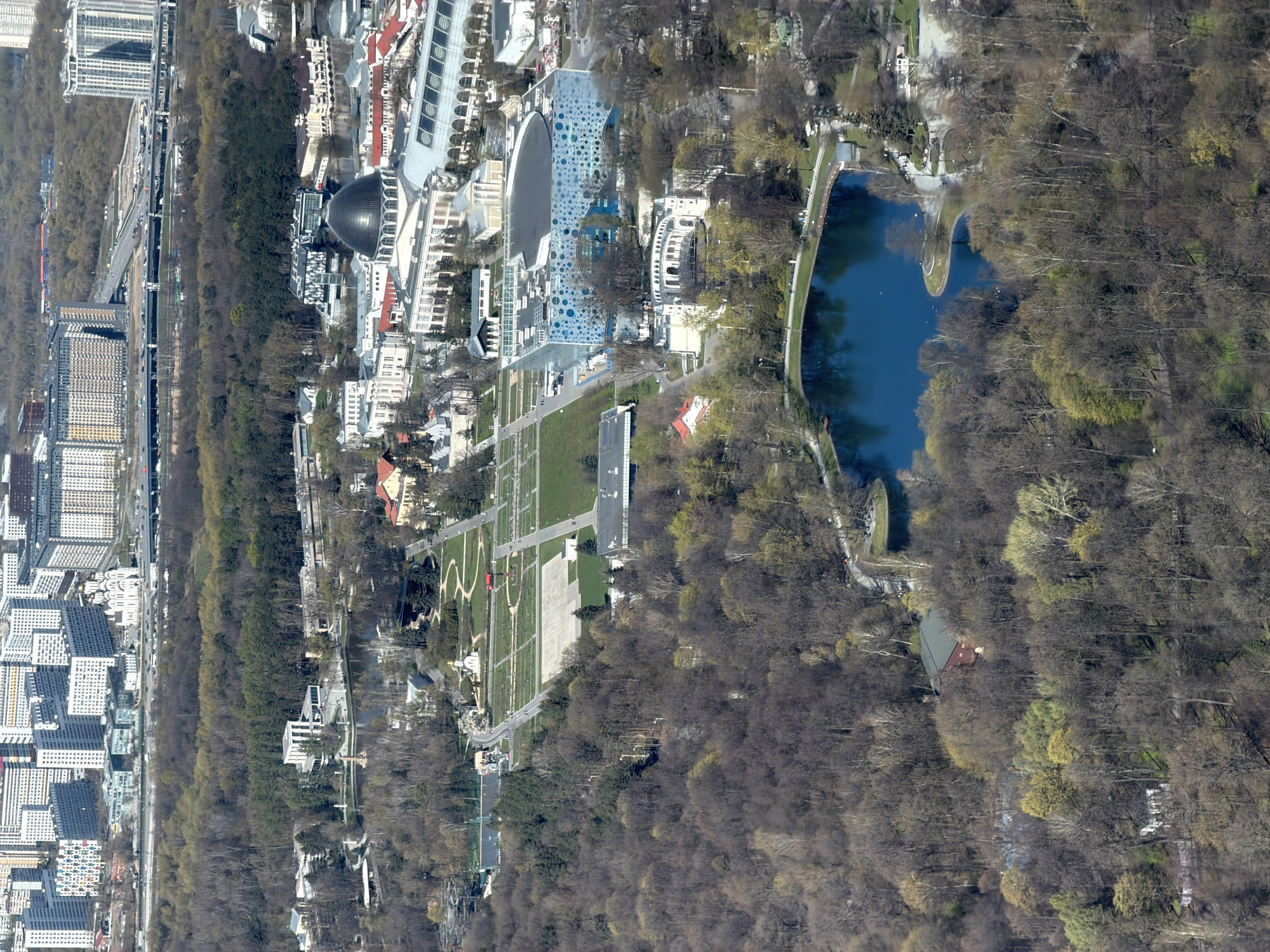 The image shows an aerial view of a park and cityscape in Khimki, Russia. To the left, modern apartment buildings and a highway run along a tree line. Adjacent to the trees is a large park, featuring an oval-shaped building with a domed roof and other modern structures, including a building with a distinctive blue, spotted facade. Further into the park is a long, narrow pond surrounded by trees. Lawns and pathways are visible, suggesting recreational areas within the park. The surrounding landscape is dominated by bare trees, indicating it is likely late fall or early spring.