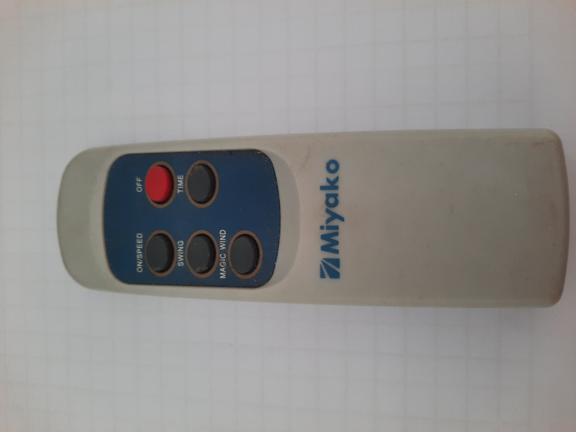 A remote control is placed on a white grid-patterned surface. The remote is light gray and has a blue panel on the upper half. On the blue panel, there are several buttons with labels: "ON/SPEED", "SWING", and "MAGIC WIND" on the left side, and "OFF" and "TIME" on the right side. The "OFF" button is red, while the others are dark gray. The brand name "Miyako" is printed in blue at the bottom of the remote. The background is a plain white surface with a faint grid pattern, suggesting it might be a desk or a table. There are no people or other objects visible in the image. The image does not provide any information about the location in Lhokseumawe, Indonesia.