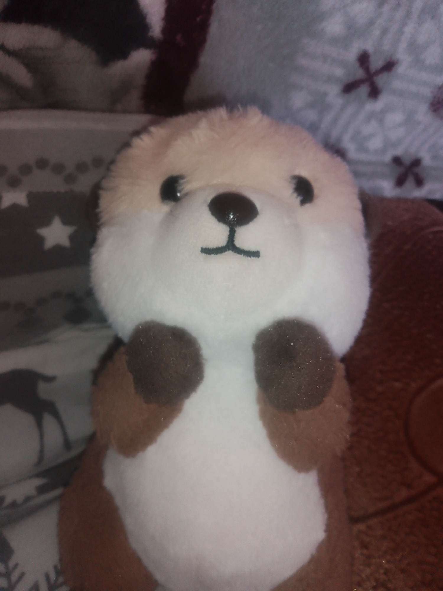 A plush toy otter sits upright on a textured brown surface, likely a sofa or chair. The otter has white fur on its belly and face, with light brown fur on its head and darker brown paws. Its eyes are black, and it has a small black nose and a stitched mouth. The background is out of focus and appears to be a patterned fabric with stars and what looks like a deer silhouette. The lighting is dim, creating a slightly blurry effect overall. The image does not contain any identifiable elements that would allow for its location to be determined in Chernivtsi, Ukraine, or any other specific city.