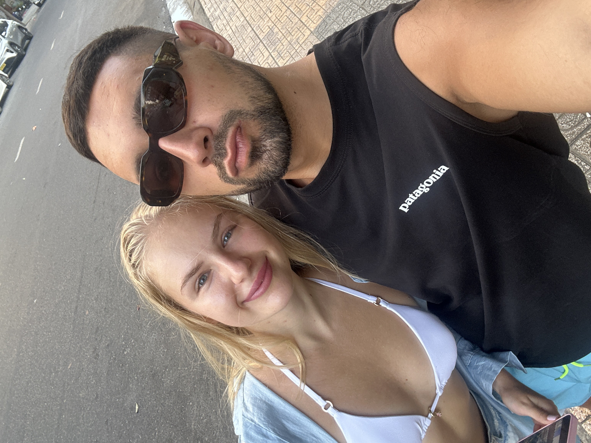 This is a selfie taken outdoors in the city of Nha Trang, Vietnam. A young couple is the main subject. The woman, on the left, has blonde hair and is wearing a white bikini top and a light blue shirt. She is smiling at the camera. The man, on the right, is wearing a black t-shirt with the "Patagonia" logo and sunglasses. He is also looking at the camera. They are standing on a sidewalk next to a street. In the background, there is a street with cars. The ground is paved with square tiles.