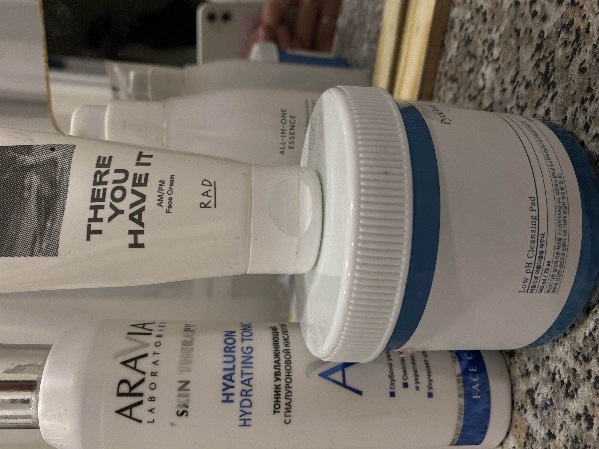 The image shows a close-up of several cosmetic products. The products include "ARAVIA LABORATORIES SKIN THERAPY HYALURON HYDRATING TONIC," "THERE YOU HAVE IT AM/PM Face Cream" with the word "RAD" written on it, and "ALL-IN-ONE ESSENCE." There is also a container of "Low pH Cleansing Pad" from the brand "Pyunkang Yul." The text on the "HYALURON HYDRATING TONIC" includes both English and Russian. The "Low pH Cleansing Pad" has Korean and English text. The background has a mottled, possibly stone-like surface. The location in the city of Kazan, Russia, is not identifiable from the image.