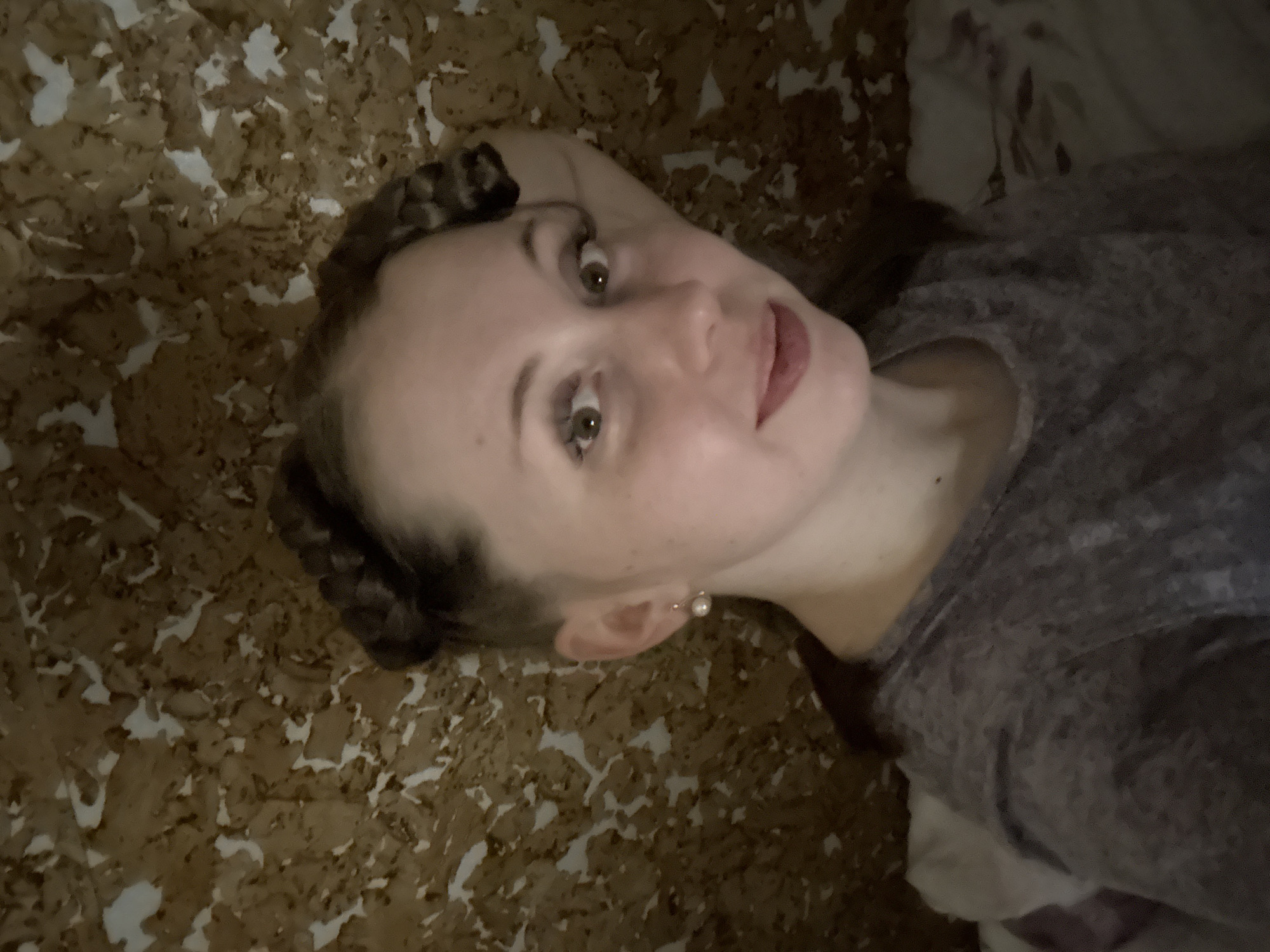 A young woman is depicted lying down, looking upwards directly at the camera. Her head is tilted, presenting a somewhat rotated perspective. She has light skin, brown eyes, and her medium brown hair is styled in two braided buns positioned on either side of her head. A single, light-colored pearl earring is visible in her right earlobe. She is wearing a grey, textured long-sleeved top. Her expression is calm, featuring a subtle smile. The background consists of a distinct textured surface, appearing as irregular patches of brown and white, reminiscent of cork board or a similar composite material. The lighting suggests an indoor environment, likely illuminated by artificial light. No visible text is present in the image.
