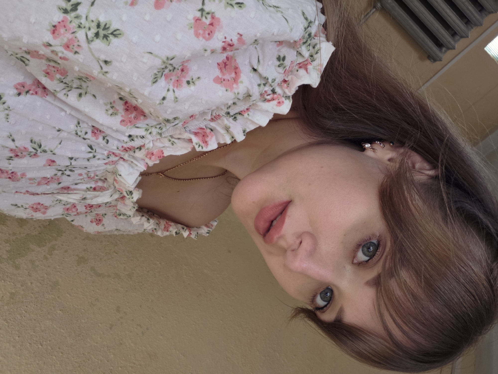An outdoor, close-up, eye-level, medium shot captures a young woman with light brown hair, styled with bangs, looking directly into the camera. She has fair skin, light blue eyes, and is wearing subtle makeup. She has a dainty gold necklace with a small pendant and a pink and white floral top with ruffled trim. The background consists of a textured, yellowish wall and what appears to be a radiator or heating element on the left side of the frame, partially obscured by the woman's hair and clothing. The lighting suggests it is daytime, with soft natural light illuminating the subject. The image does not contain any discernible elements to indicate its specific location within Chaikovsky, Russia.