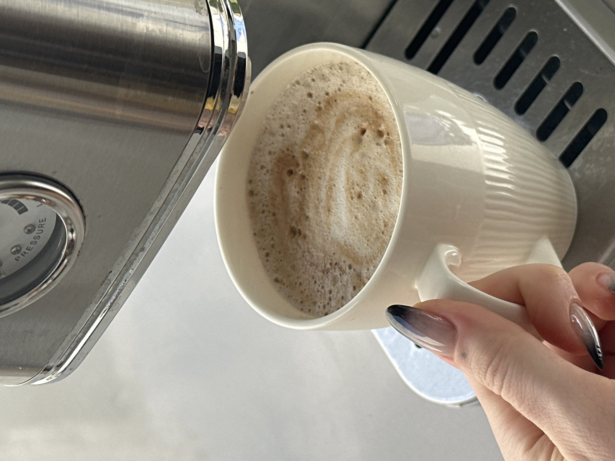 A close-up shot shows a hand with dark-tipped, ombre-style manicured nails holding a white ceramic mug filled with coffee and crema. The mug sits on a metal grate, part of a coffee machine. The machine's stainless steel surface and a pressure gauge are visible at the top of the frame. The scene appears to be indoors, with a light-colored, possibly concrete surface in the background. The specific location in North Kuta, Indonesia, is not discernible from the image.