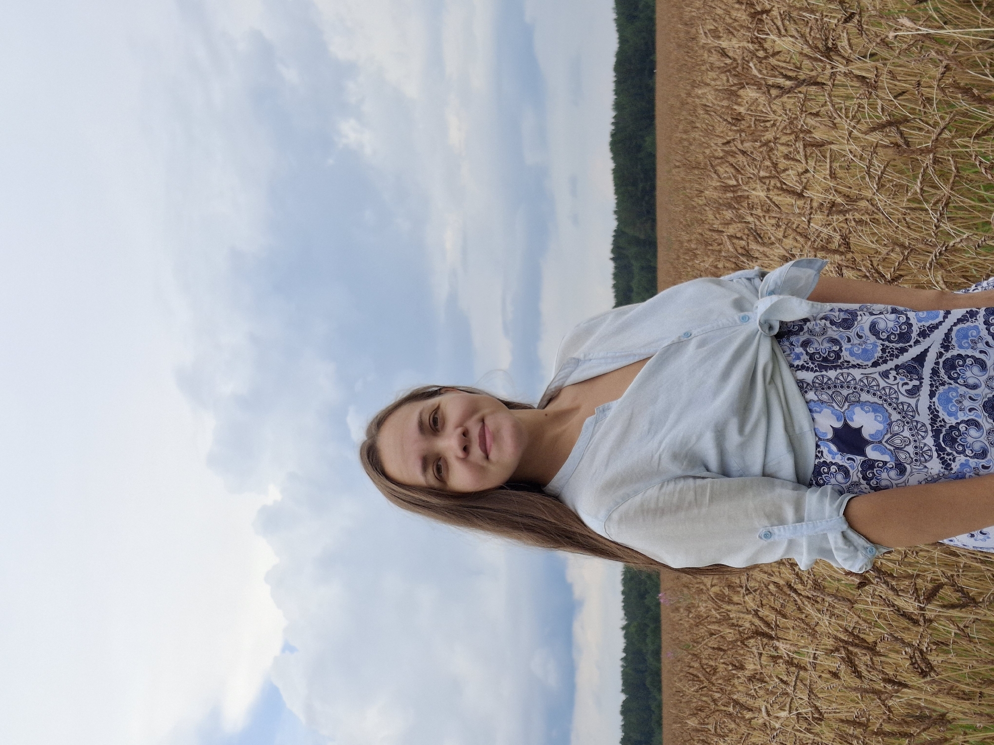 A medium shot shows a woman standing in a wheat field, with a forest line and cloudy sky in the background. The woman has long brown hair and is wearing a light blue, unbuttoned shirt tied at her waist, over a patterned blue and white skirt. Her gaze is directed at the camera. The wheat stalks are dry and golden, filling the foreground and midground. The forest forms a dark green horizontal band against the horizon. The sky is overcast with large, light gray clouds. The location is identified as Plishkino, Russia.