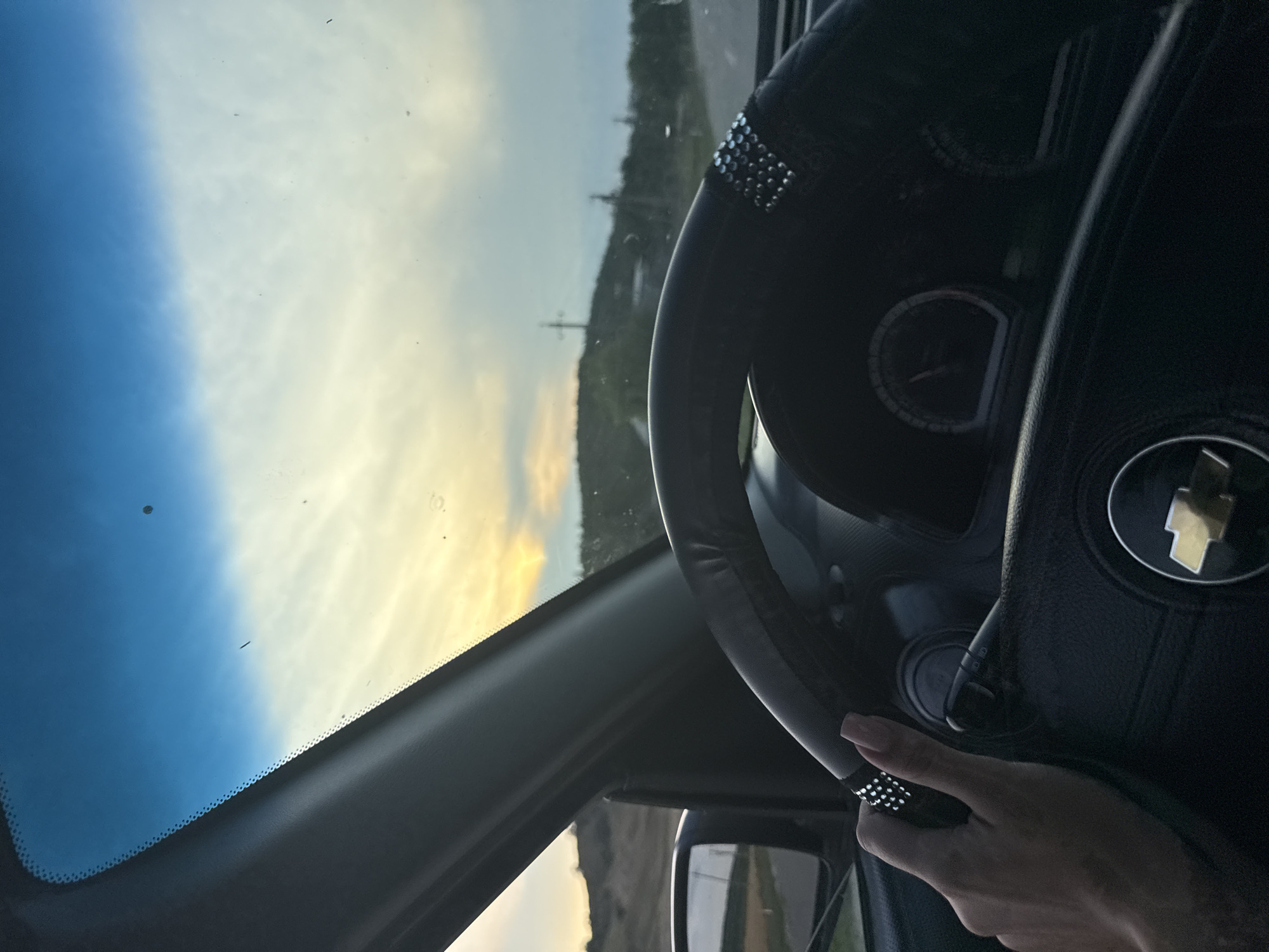 The image shows the interior of a car, viewed from the driver's perspective. A hand is visible on the steering wheel, which is covered in a black case adorned with silver studs. The dashboard and steering wheel are dark, with a Chevrolet logo visible on the latter. Through the windshield, a sunset is seen with a gradient of blue and yellow sky. The landscape outside appears rural, with hills and scattered vegetation. The location is specified as the city of Sadaclia, Moldova, though this information cannot be visually confirmed from the image itself. The overall impression is that of a person driving at dusk in a semi-rural area.