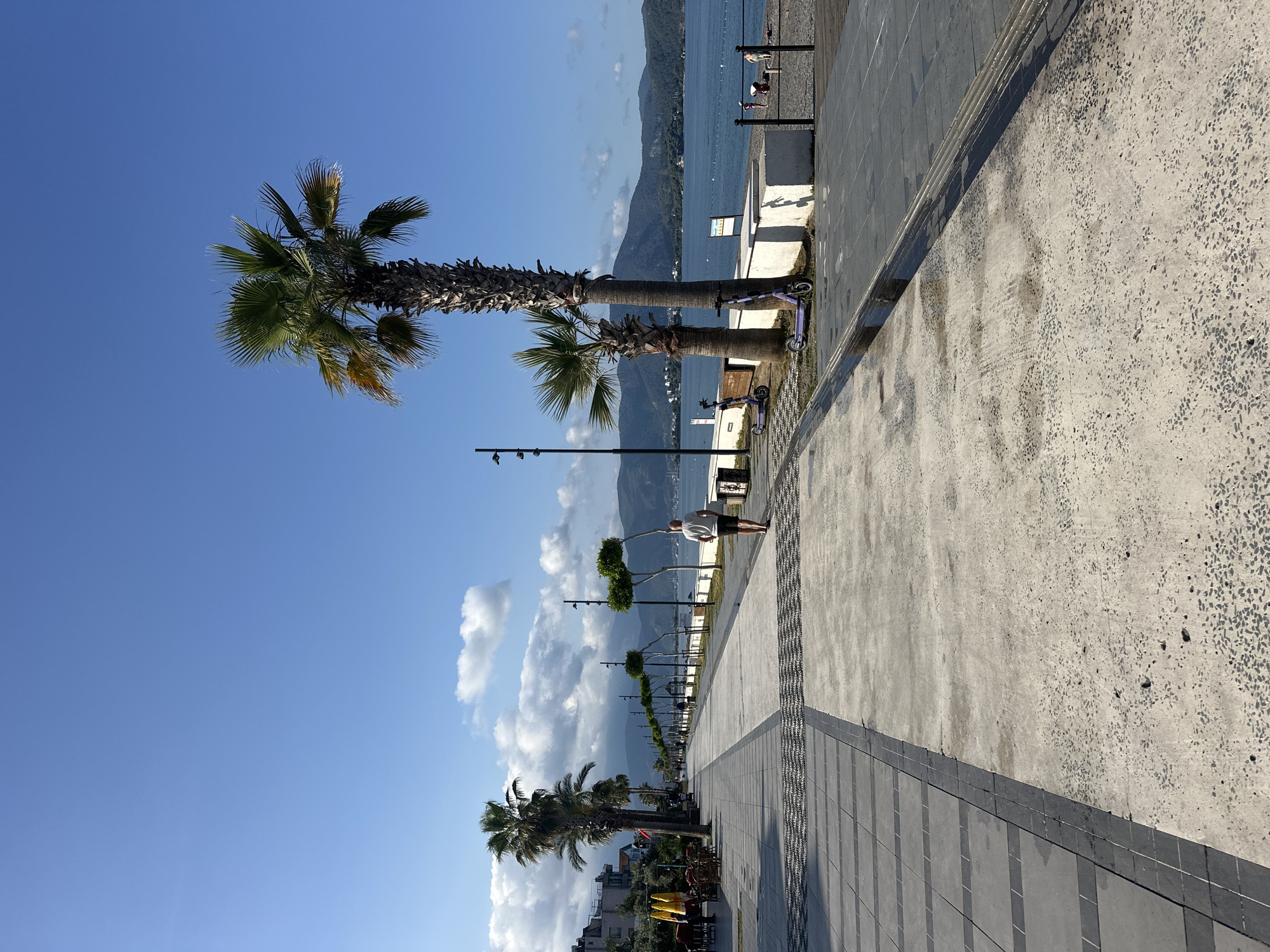 Here is a description of the image:
The photograph shows a waterfront promenade in Fethiye, Turkey. The promenade runs along the sea and is lined with palm trees and decorative trees. In the distance, mountains are visible. The sky is blue with scattered clouds.
A person is standing on the promenade. There are also electric scooters parked along the edge of the promenade. The promenade is paved with concrete and gray tiles.