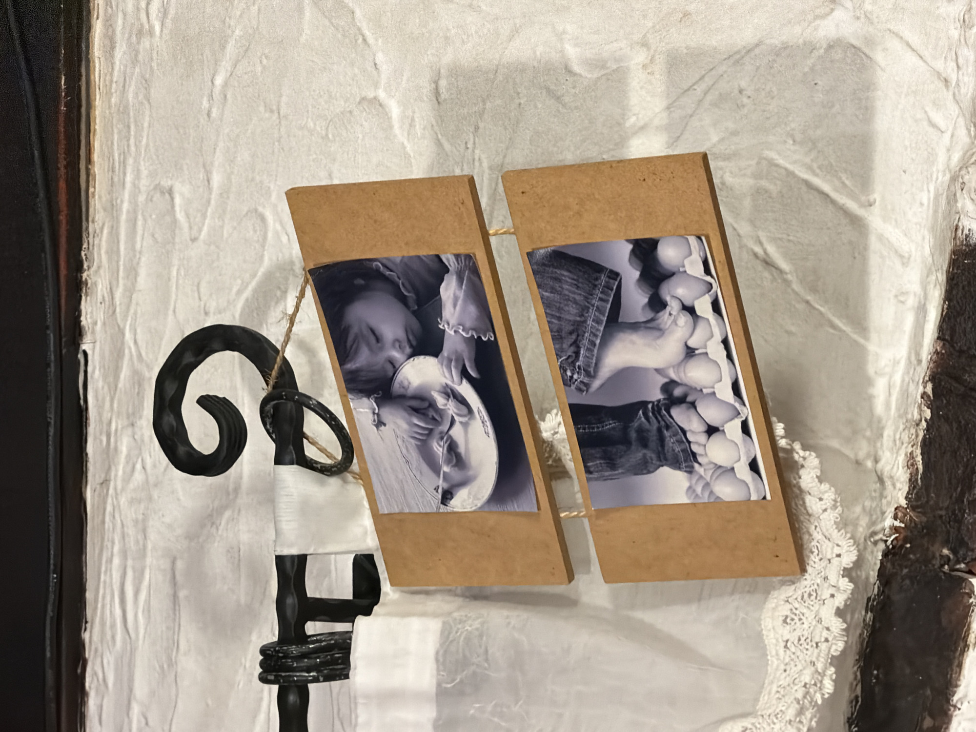The image depicts a black and white photograph of a child eating from a plate, and another photograph of a child's feet, each housed in a wooden frame and hung side-by-side on what appears to be a textured wall. A black wrought-iron decorative element is attached to the wall next to the photographs. A white cloth with lace trim is draped beneath the photographs and ironwork. A dark, vertical structure is visible on the left and right edges of the frame. There is no information available that indicate the location to be in Varna, Bulgaria.