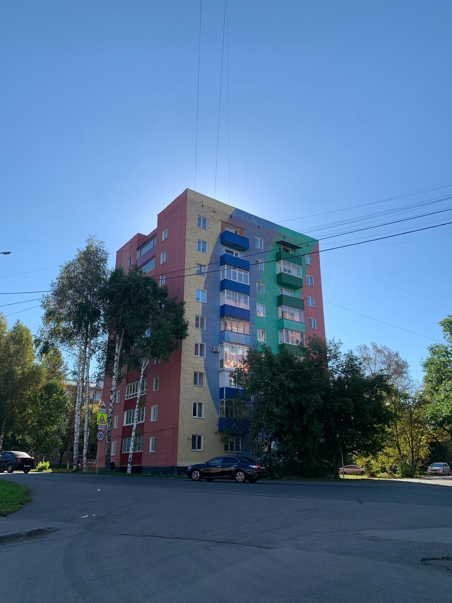 The image shows a multi-story residential building in Prokopyevsk, Russia, painted in bright, alternating sections of pink, yellow, blue, and green. The building has several floors with numerous windows and balconies, some of which are enclosed with glass and others are open. Tall birch trees with white trunks and green foliage stand in front of the building, partially obscuring some of its colorful facade. A dark colored sedan is parked on the right side of the street in front of the building, and another dark colored car is seen further down the road. On the left side of the street, a black sedan is driving away from the camera. A traffic sign with the number 20 is visible near the sidewalk. The sky is clear and blue, with a few power lines running across the top of the frame. The overall impression is of a sunny day in an urban environment with a visually striking, colorful building.