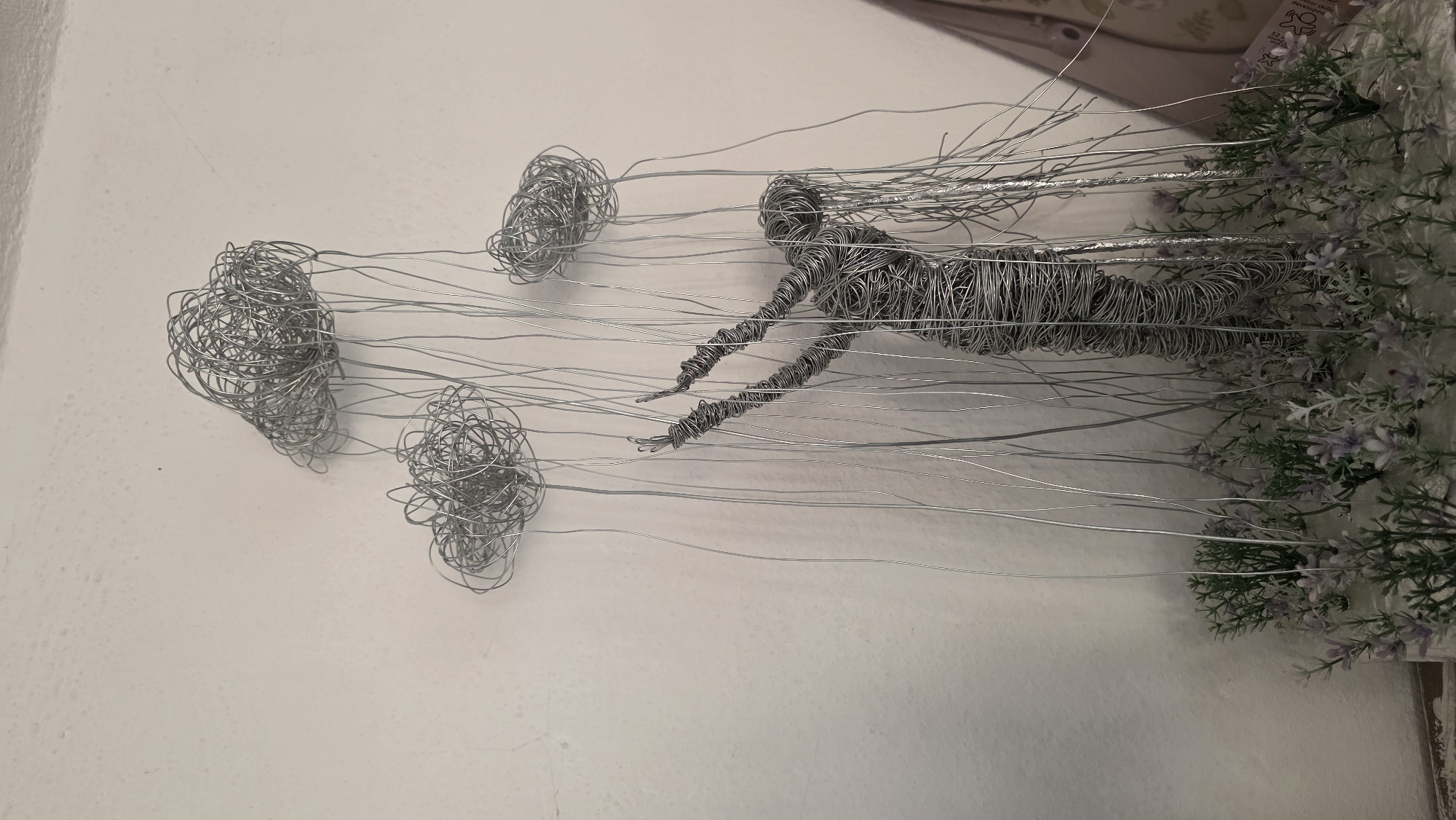 A wire sculpture of a swimmer with two clusters of wire representing bubbles is attached to a wall. A bed of artificial flowers is positioned at the right edge of the sculpture, appearing to be the swimmer's origin or end point. The background is a plain white wall. The location of the image is unknown, so I am unable to provide information about the city of Blagoevgrad, Bulgaria.