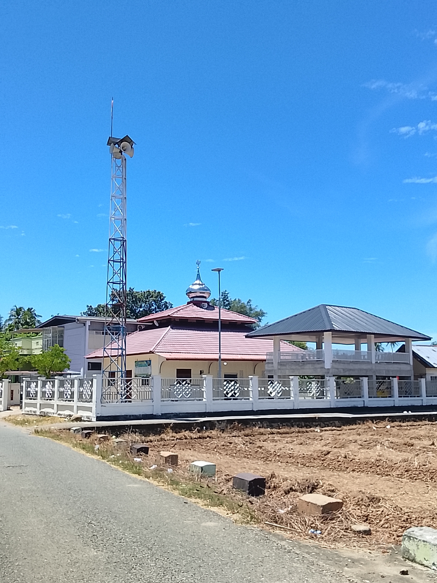 The image depicts a clear, sunny day in Rhieng Krueng, Indonesia. In the foreground, a paved road leads the eye towards a white fence with ornate metalwork. Beyond the fence, a mosque with a red-tiled roof and a silver dome is visible. To the left of the mosque, a tall, silver metal tower with multiple speakers stands against the bright blue sky. Further to the right, a covered structure with a dark, sloped roof is supported by white pillars, appearing to be an open-air pavilion or seating area. The ground behind the fence is uneven and appears to be recently disturbed or under construction, with scattered brown soil and some debris. Lush green trees are present in the background, suggesting a tropical or subtropical climate. There are no people visible in the image. The scene is tranquil and static, capturing a moment of everyday life in the Indonesian town.