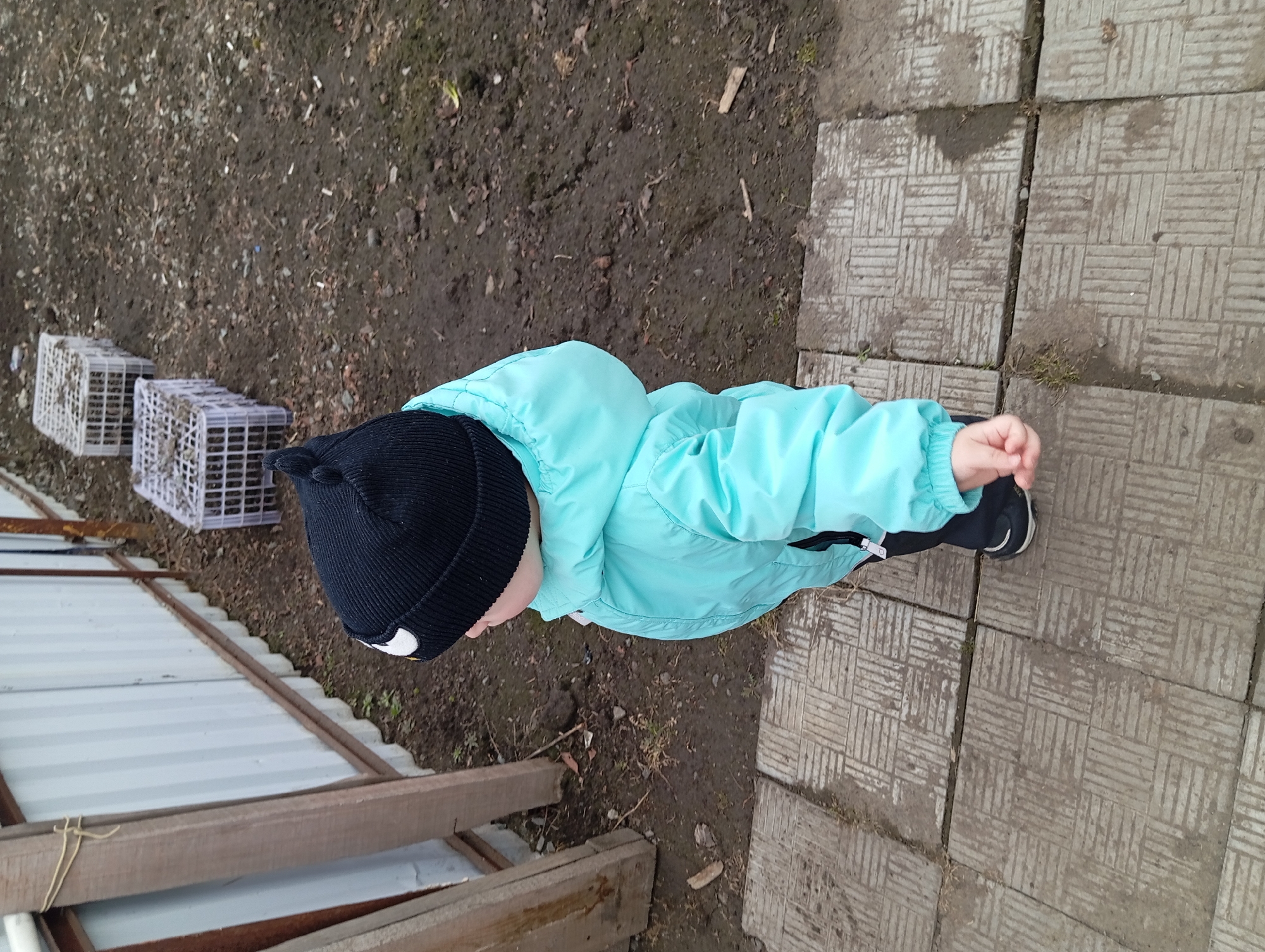 A child wearing a black beanie and light blue jacket stands on patterned concrete pavers. Adjacent to the pavers is bare, dark soil with scattered debris. To the left, near the soil, are several white plastic crates and a section of corrugated metal fencing with wooden supports. The scene appears to be outdoors in Yurgamysh, Russia.