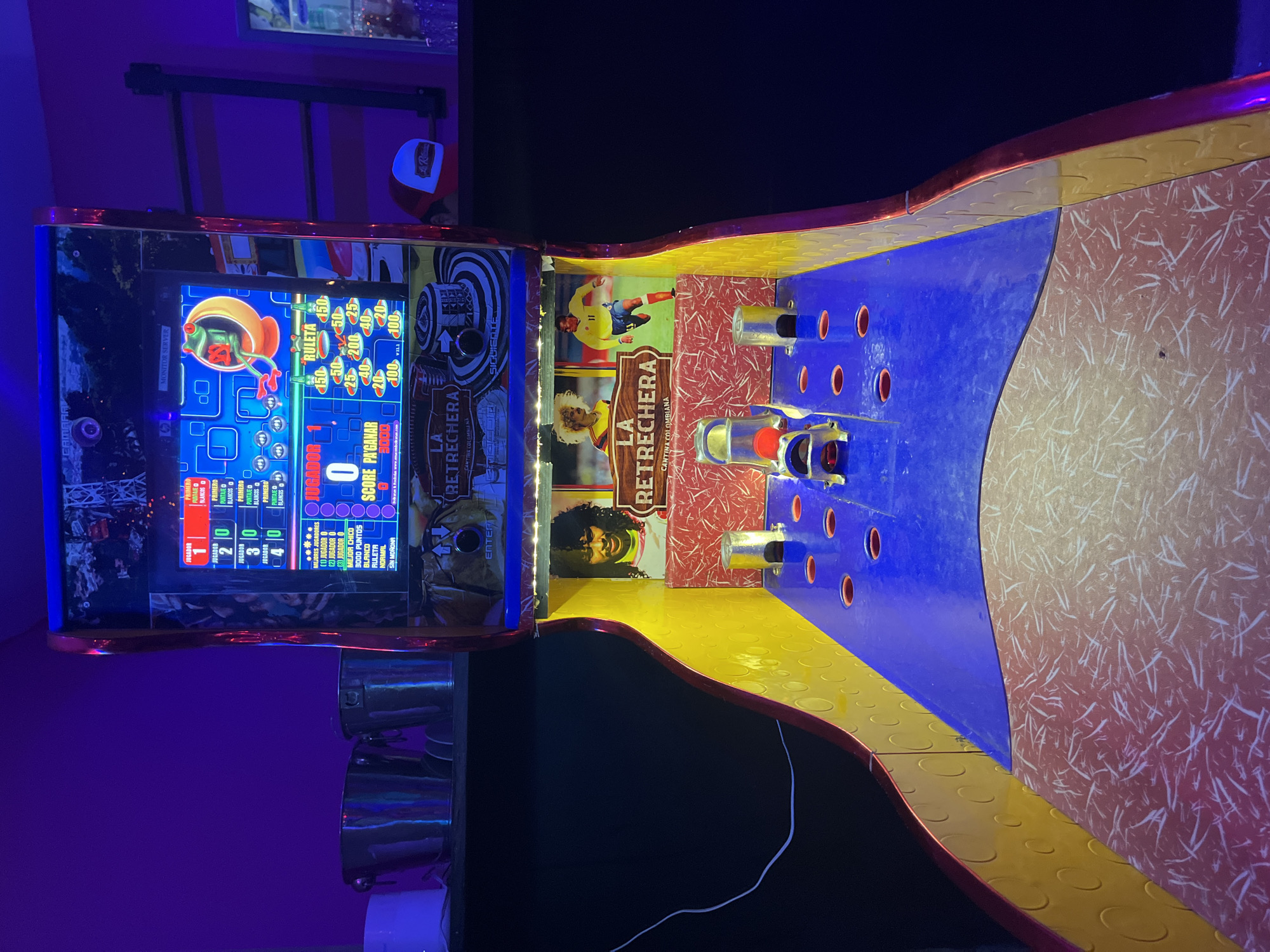 The image shows an angled overhead shot of an arcade game, possibly located in Buenos Aires, Argentina, given the game's branding with "LA RETRECHERA CANTINA COLOMBIANA," suggesting a Colombian theme or connection. The game features a monitor screen displaying game-related graphics, including a frog image and various point values. Below the screen are slots with lit buttons and displays indicating player scores. The game's physical play area is a track-like structure with red, yellow, and blue sections, featuring several holes and cylindrical targets. A red game piece sits on the blue section. The surroundings are dimly lit with purple ambient light visible on a wall to the left. A red baseball cap rests atop the game.