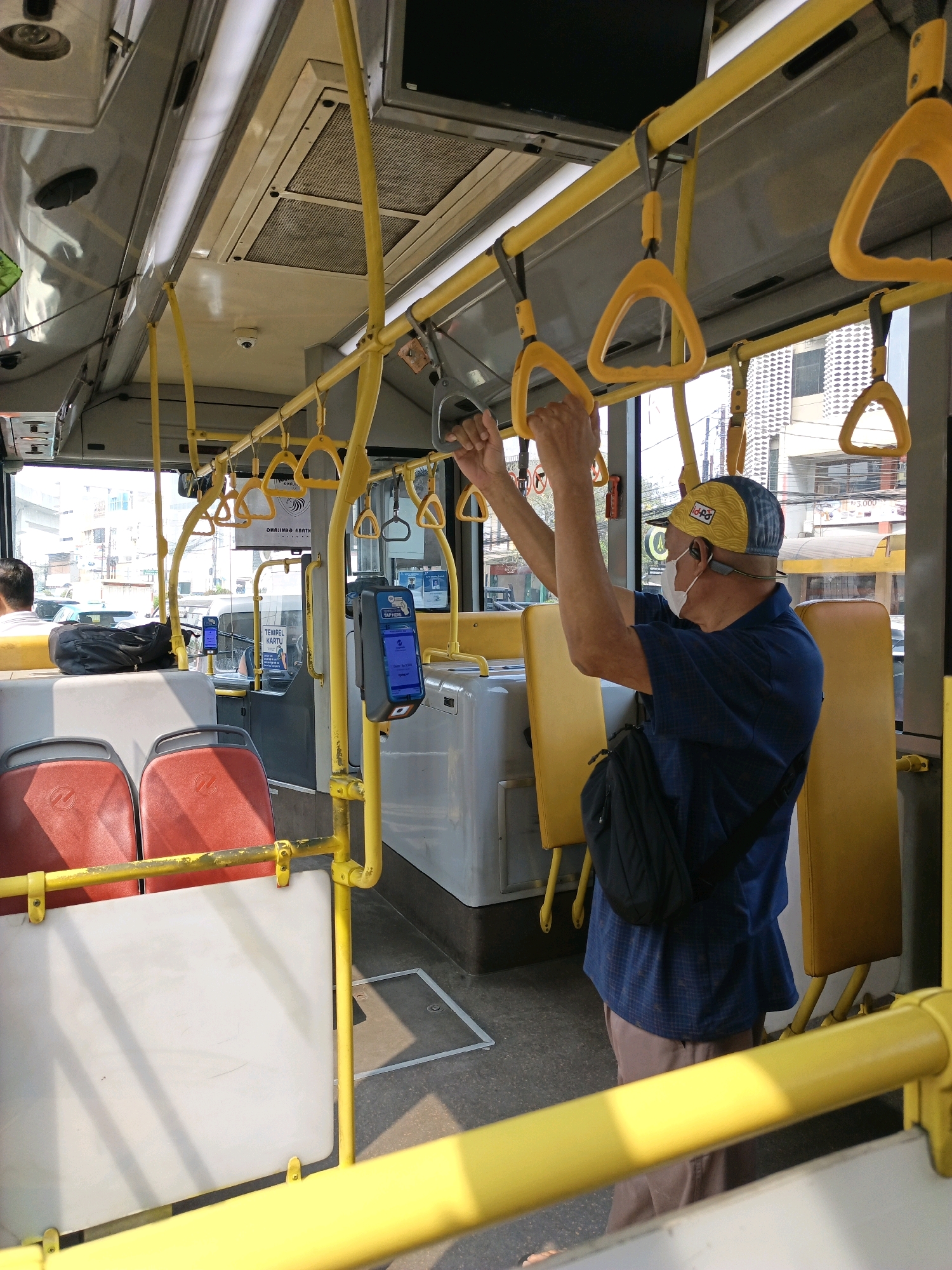 The image shows the interior of a public bus in South Jakarta, Indonesia. A man wearing a blue checkered shirt, brown shorts, a yellow and blue baseball cap, and a white face mask is standing and holding onto yellow handrails. He has a black messenger bag slung across his chest. The bus is equipped with numerous yellow handrails and vertical poles for passengers to hold onto. Red seats are visible in the foreground, and yellow seats are further back. A card reader machine is mounted on a pole. Sunlight streams in through the windows, casting shadows on the floor and seats. Outside the bus, buildings and other vehicles are visible.