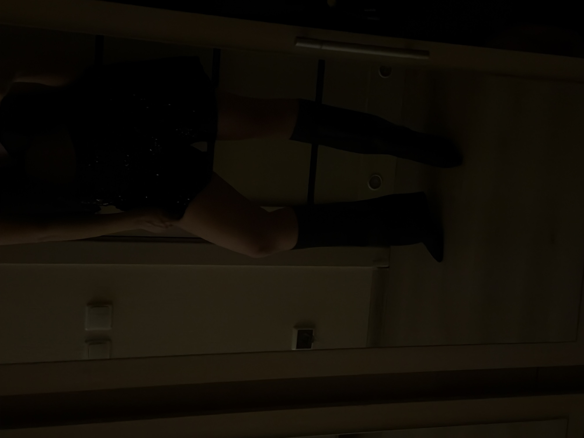 The image depicts a person wearing a dark dress and black knee-high boots. The person is positioned against a light-colored wall, potentially a cabinet or storage unit, which features a metallic handle and what appears to be light switches. The lighting is dim, obscuring details. There is no information in the image to indicate it was taken in Izhevsk, Russia.
