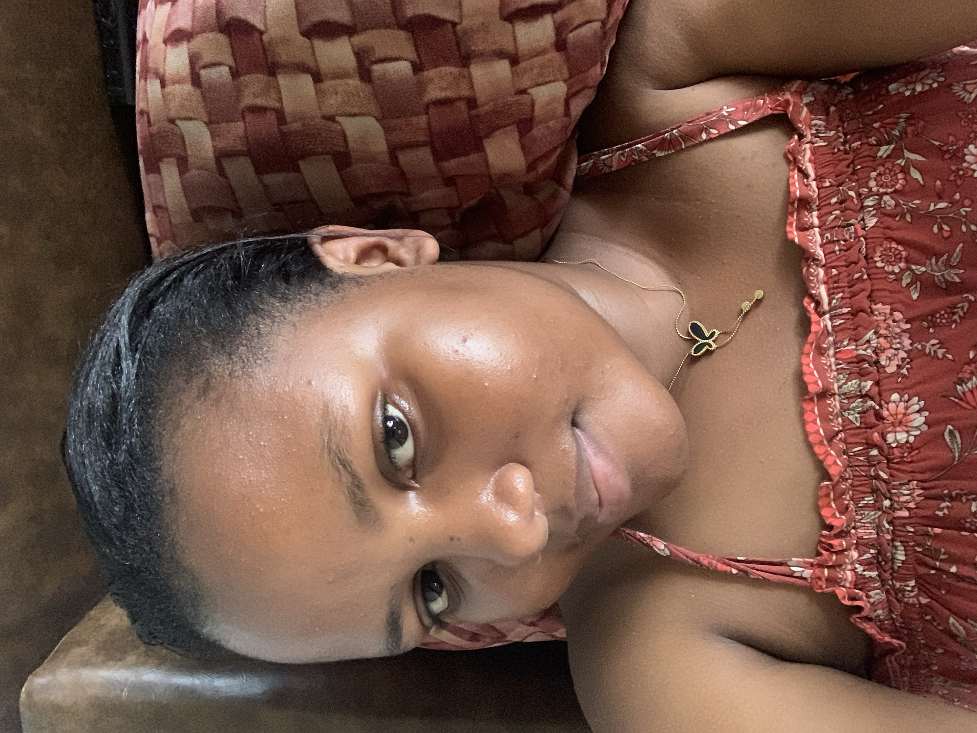 A close-up, tilted-angle shot shows a woman with dark hair and brown skin lying down. She is wearing a red floral-patterned top with thin straps and a gold necklace with a butterfly pendant. Her head is resting on a brown and red patterned pillow. The background is a brown surface. The location cannot be determined from the image.