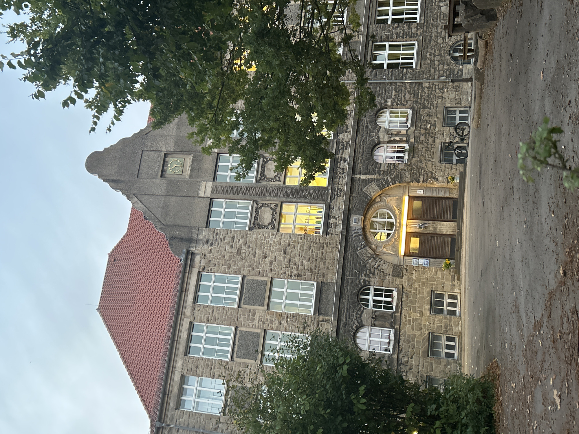 This is a medium shot photograph of the front of a stone building, likely a school, with a red tile roof. The building has numerous white-framed windows of various sizes, some with visible glass panes and others with a grid-like structure. On the upper facade, a round clock with Roman numerals is visible. There is a large arched entrance in the center of the building with double wooden doors. To the left of the entrance, a green tree with dense foliage partially obscures the building. To the right of the entrance, another tree with similar foliage is visible, also partially obstructing the view. The ground in front of the building is a paved area with scattered leaves. A bicycle is parked to the left of the entrance, and a wooden bench and rocks are situated on the right side of the frame, beyond the paved area. The sky is overcast. The building is identified as the Christian-Dietrich-Grabbe-Gymnasium located in Detmold, Germany.