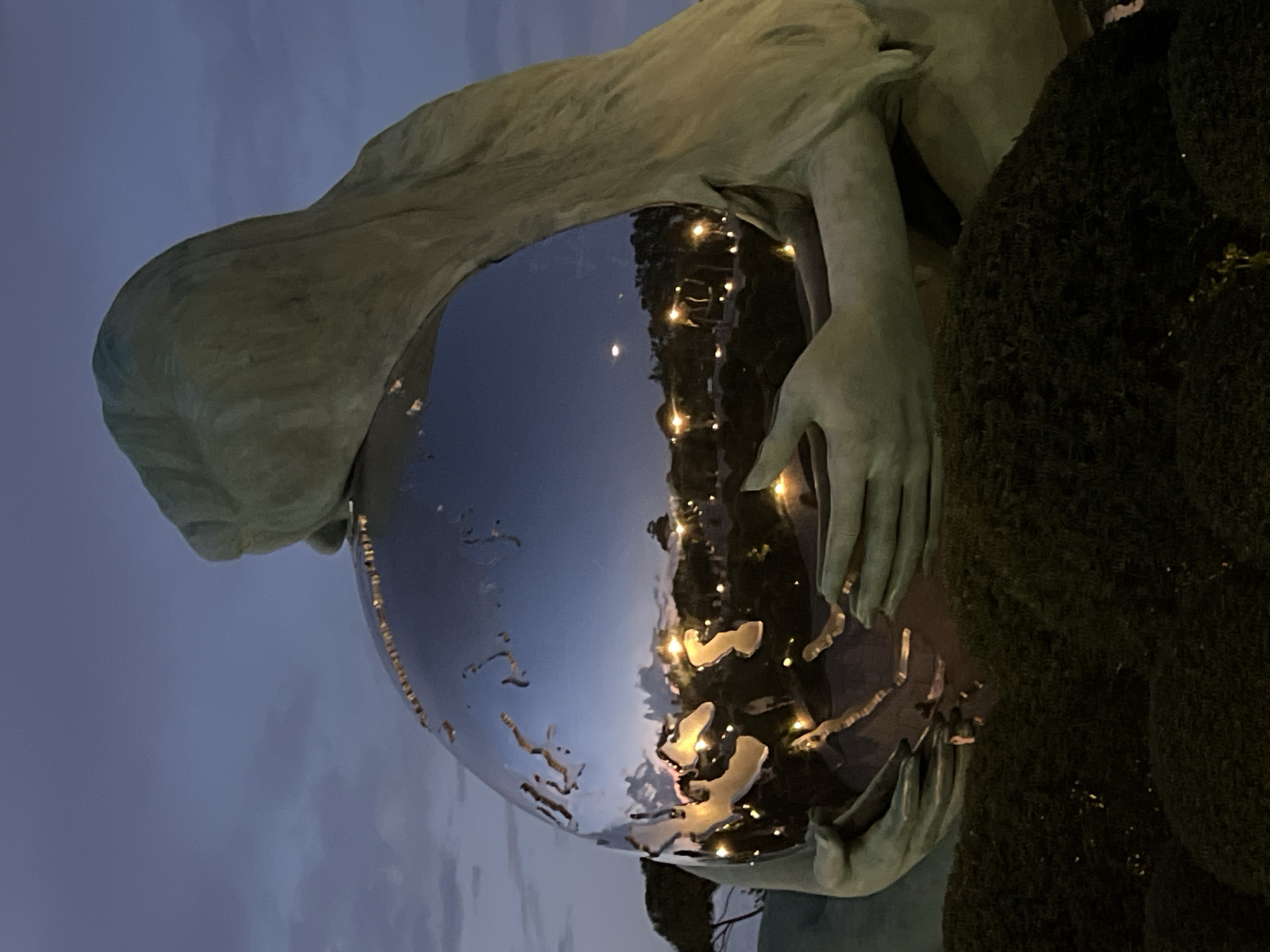 This is a close-up, rotated image of a sculpture at dusk. The sculpture appears to be made of a light-colored material, possibly bronze or stone, and depicts a figure holding a large, reflective sphere. The sphere, which resembles a globe, reflects the surrounding environment, including a dark sky with hints of clouds and a line of illuminated lights that could be from buildings or streetlamps. The figure's hands are gently cupping the sphere, and part of what looks like a draped garment is visible. To the right of the sphere, there's a textured, dark green surface, likely a bush or other foliage, also illuminated by the distant lights. The overall mood is contemplative and serene, enhanced by the twilight setting. The image does not contain any visible text. The location context is Krasnodar, Russia.